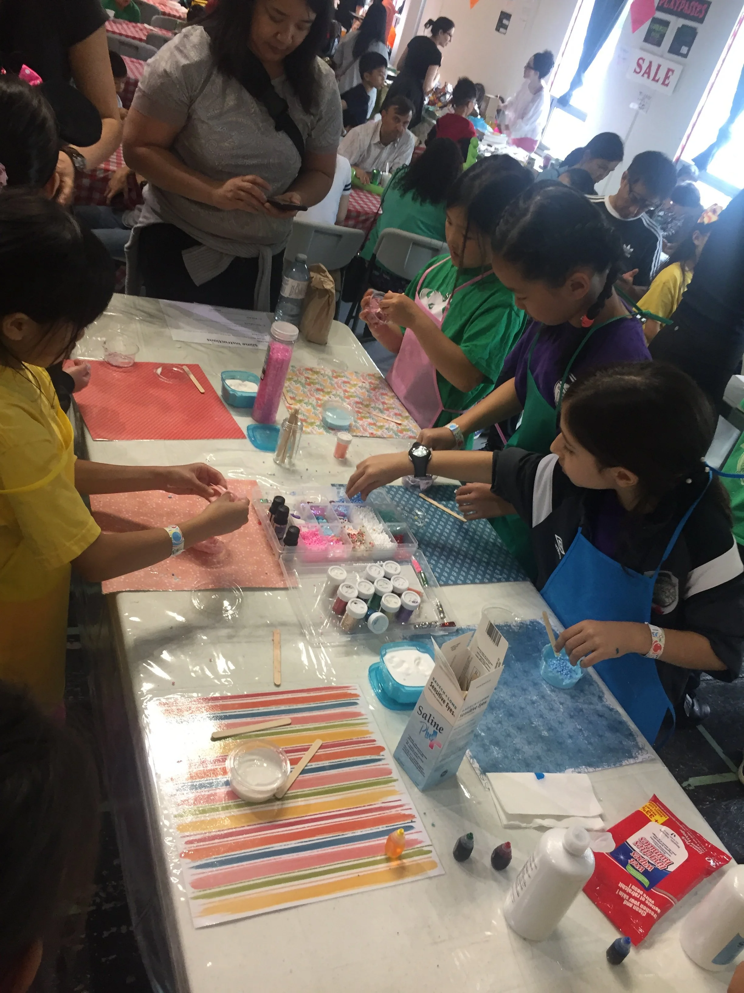 Children and adults gathered around a table engaging in a craft activity involving colorful powders and water, with various craft supplies like bottles, spoons, and containers on the table, in a busy indoor setting.