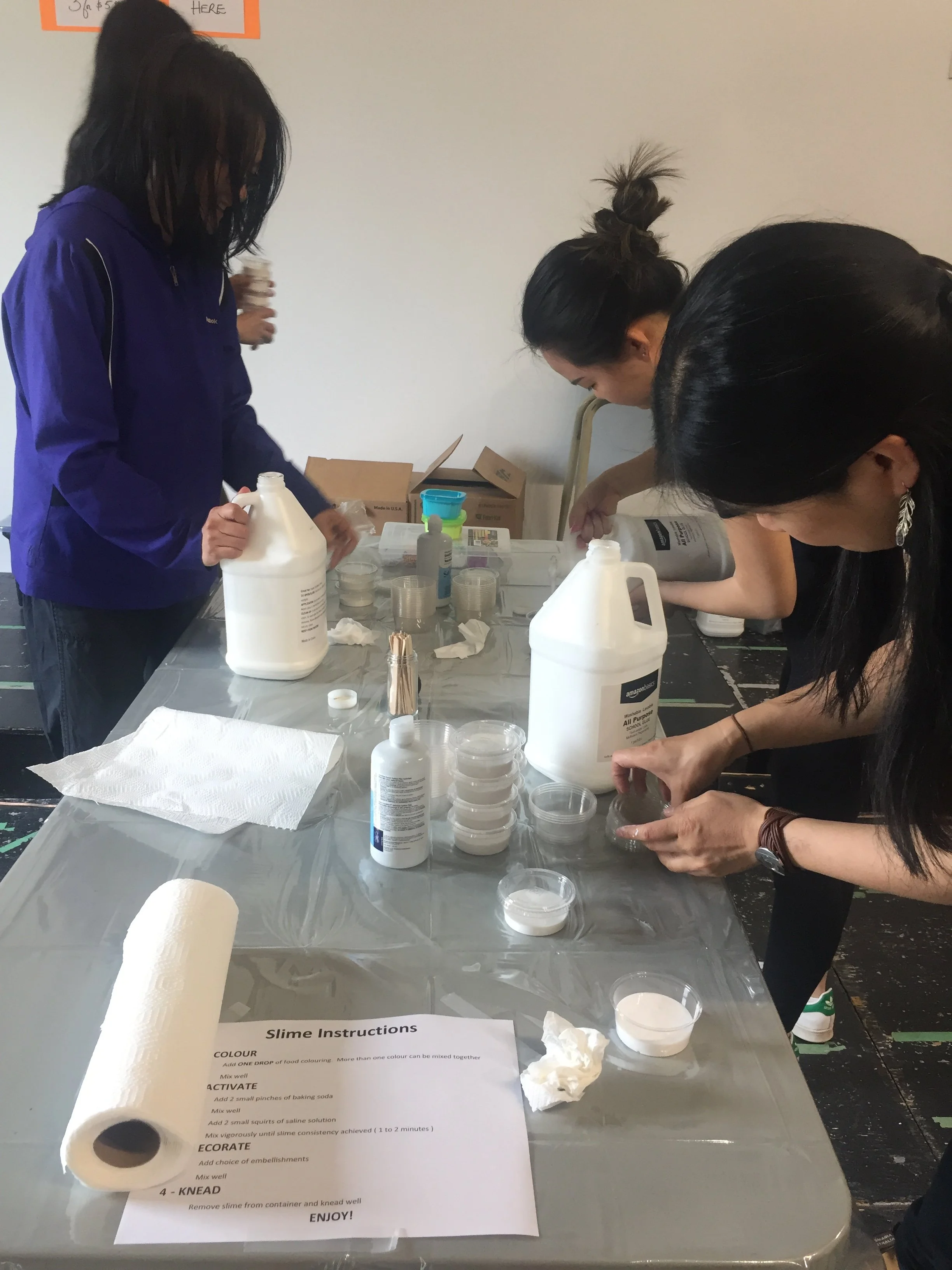 Three women are working on a slime-making project at a table covered with plastic, with bottles, containers, and instructions visible.
