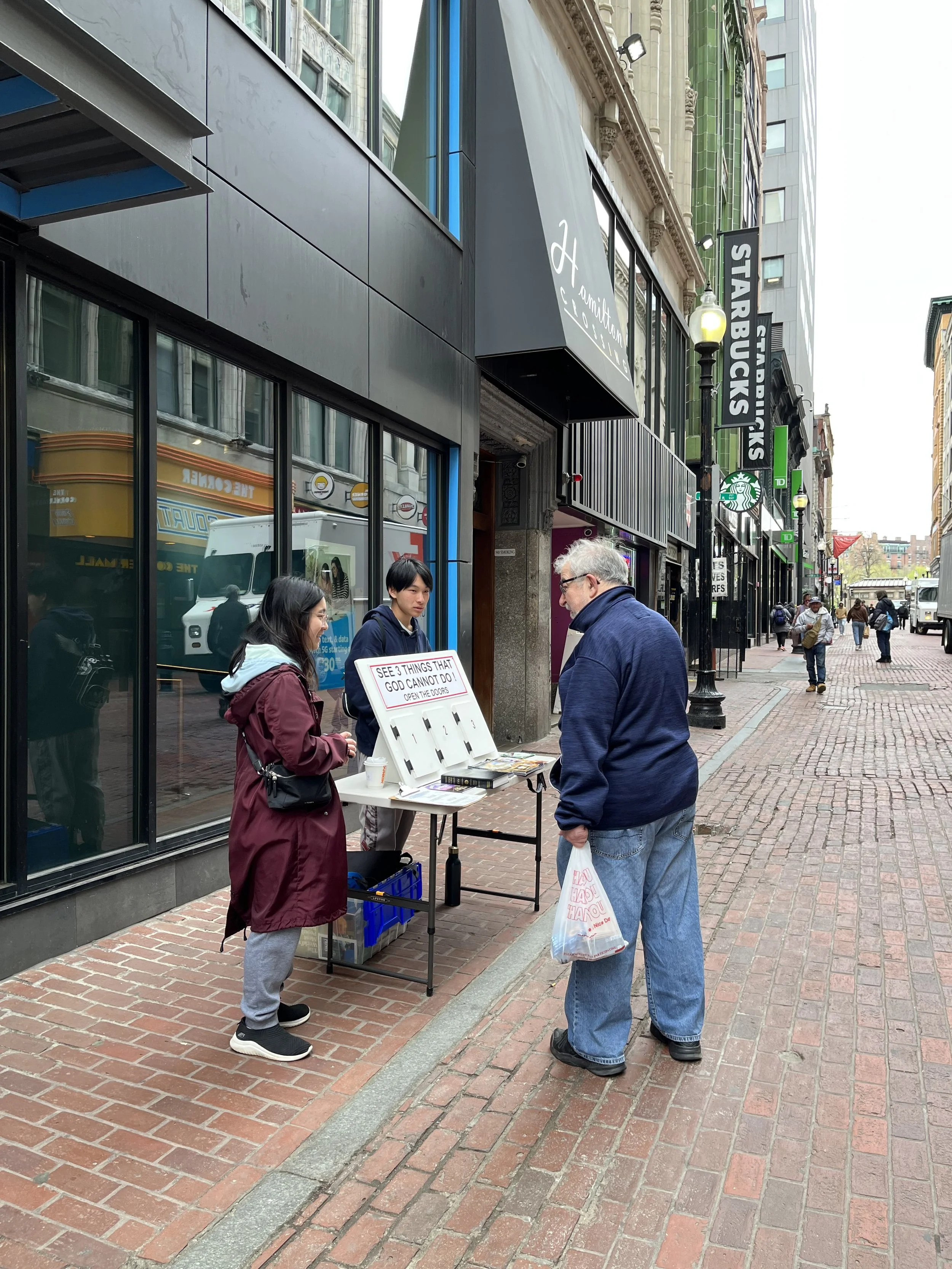 Day 4 - Mission team members speaking to an individual on a street with a display board that says "3 Things God Cannot Do"