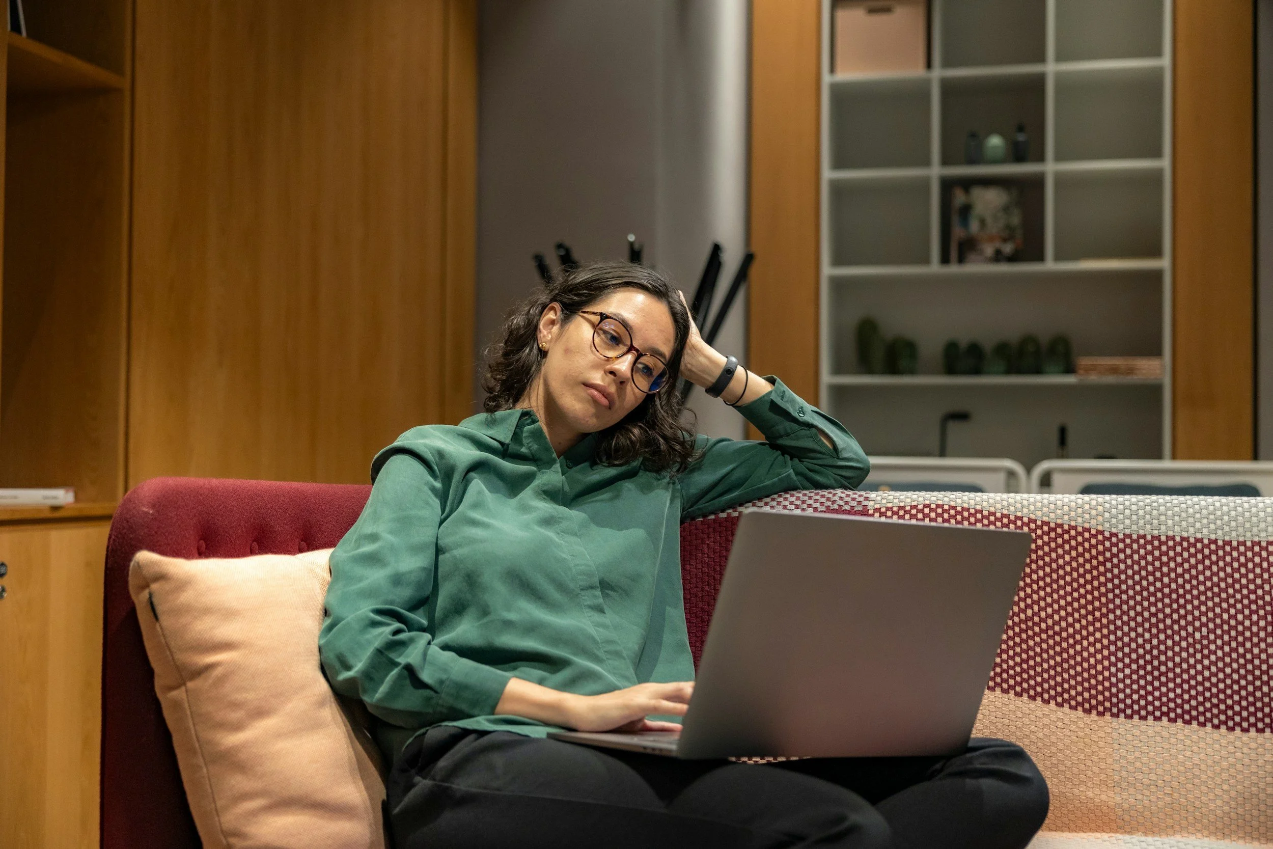 A woman with glasses and dark curly hair sitting on a red chair at a table, working on a laptop with a focused expression, in a room with wooden walls and a shelving unit in the background.