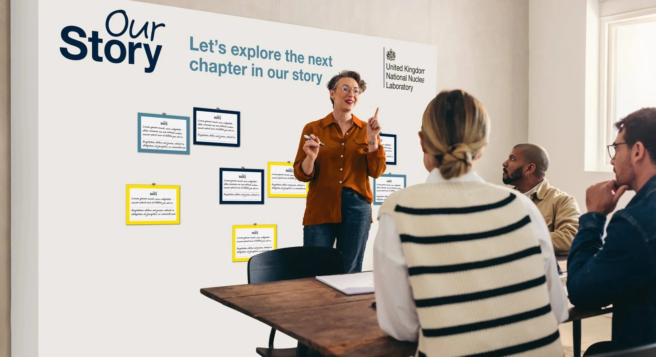A woman in an orange shirt speaking to a group of four seated people in a classroom or meeting room. Behind her, there is a large whiteboard with the words 'Our story' and 'Let's explore the next chapter in our story' along with framed papers attached to the board. The room has a window letting in natural light.