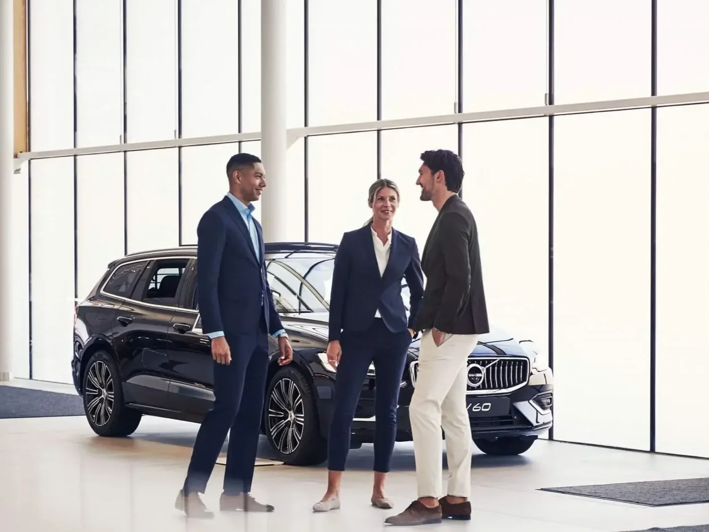 Three businesspeople talking and smiling inside a car dealership with a black Volvo SUV in the background.