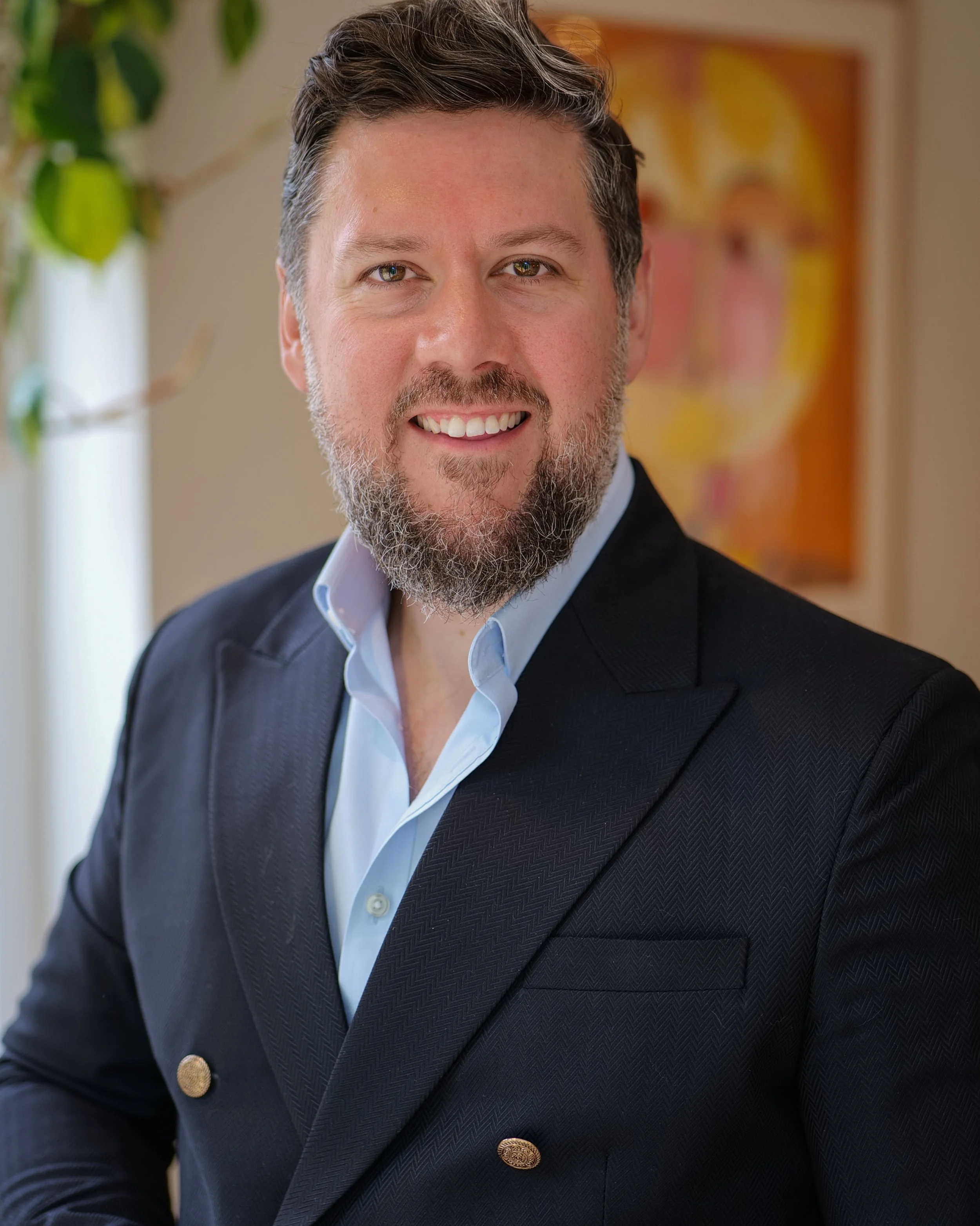 Close-up of a smiling man with dark hair, beard, and light skin, wearing a dark blazer and light shirt, in a well-lit room with a green plant and colorful abstract artwork in the background.