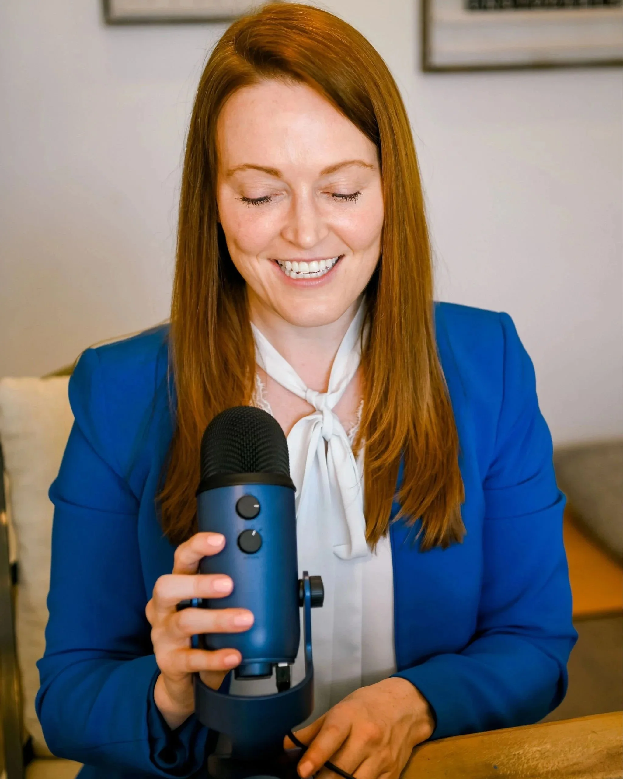 A woman with long red hair wearing a blue blazer and white blouse smiling while holding a blue microphone in her right hand, sitting at a wooden table.