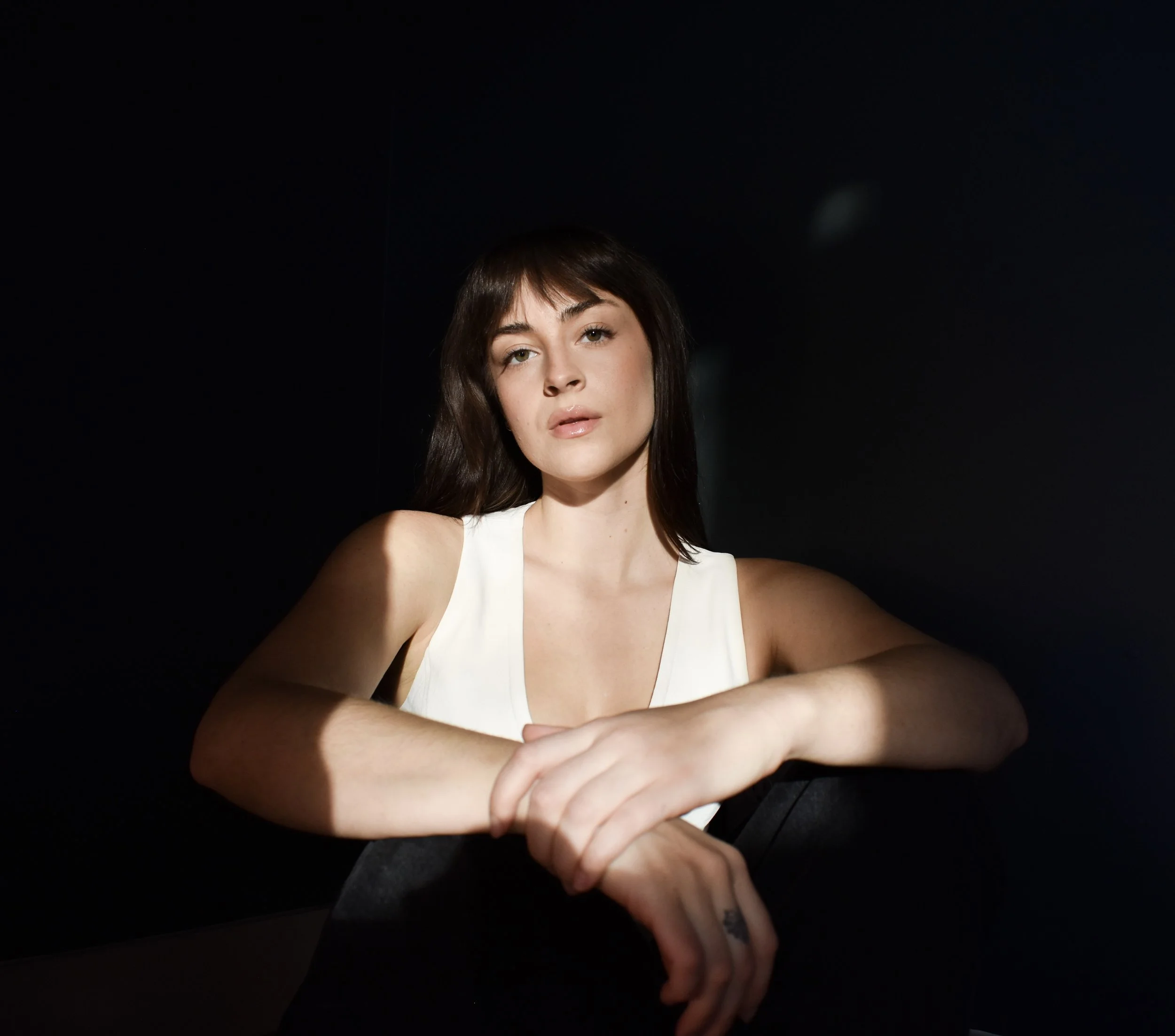 A young woman with dark hair and fair skin sitting at a table with her arms crossed on the surface, in front of a black background.