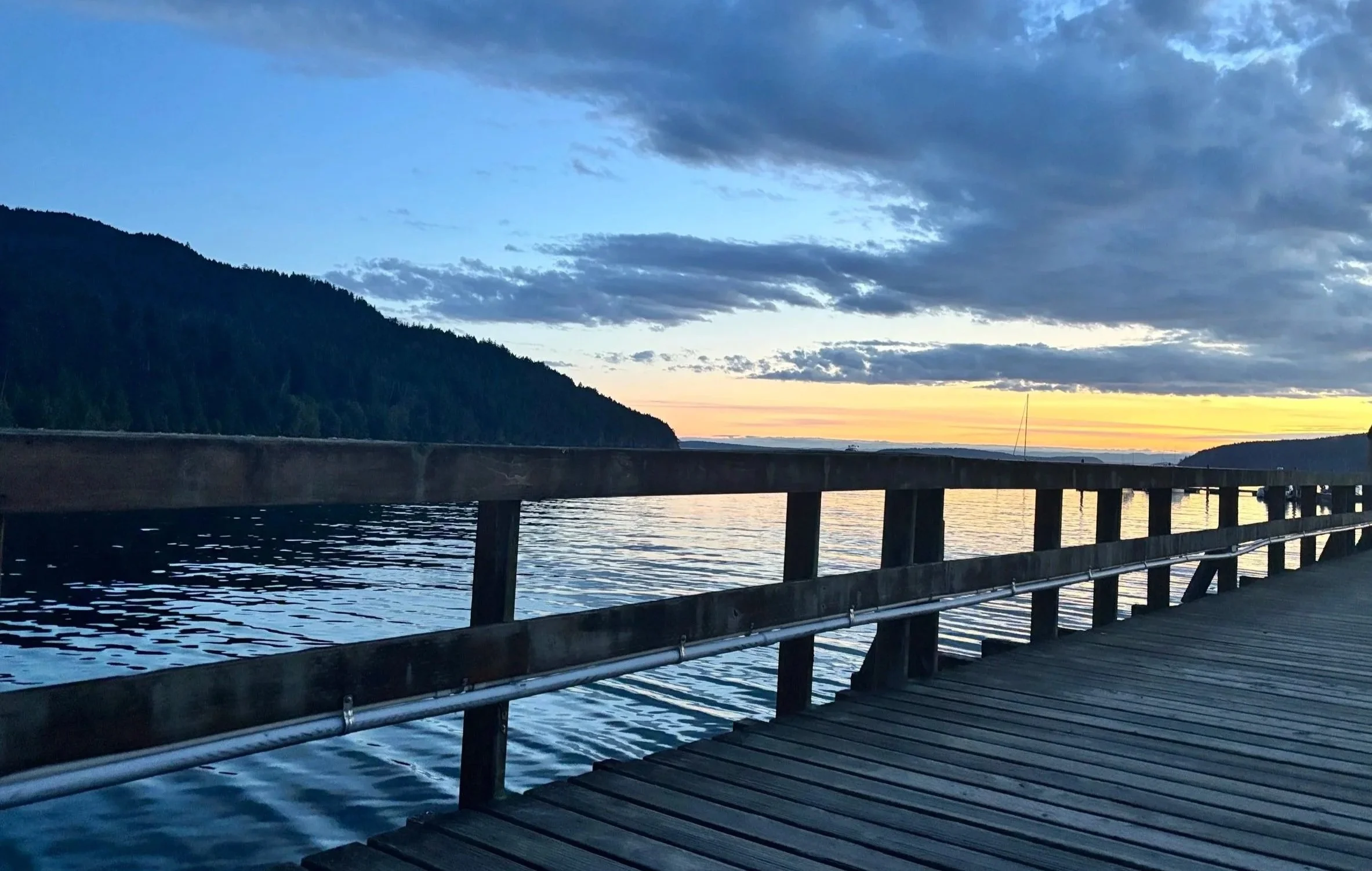 A wooden pier extends over a body of water with hills covered in trees in the background. The sky is partly cloudy with the sun setting on the horizon, casting a warm glow.