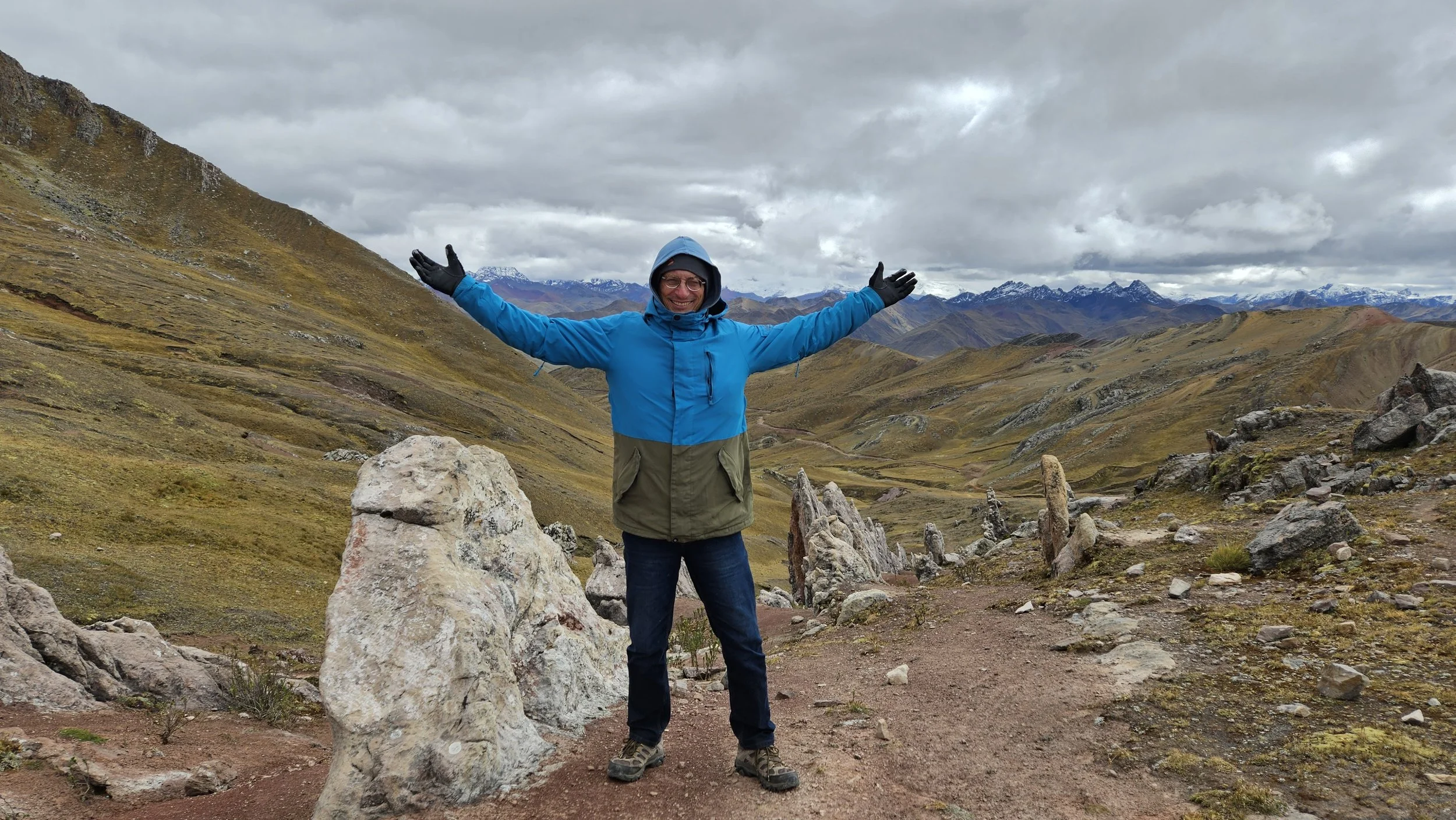 A person in a blue jacket and black gloves standing with arms outstretched in a mountainous landscape with rolling hills, rocks, and snow-capped peaks in the distance under a cloudy sky.