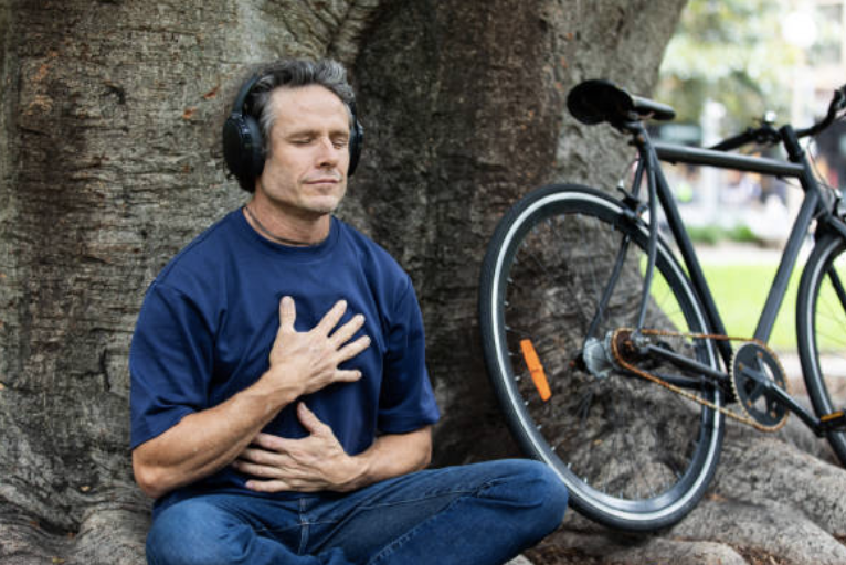 A man sitting cross-legged outdoors by a large tree, wearing headphones, with one hand on his chest and the other on his stomach, appearing to meditate or relax. A bicycle leans against the tree behind him.