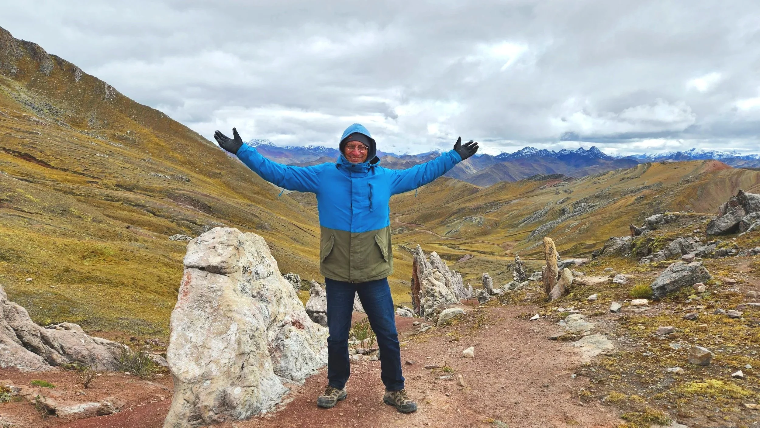 A person in a blue and green jacket with a hood, glasses, and gloves standing with arms outstretched in a mountainous landscape with rolling hills, rocky formations, and snow-capped peaks in the distance.