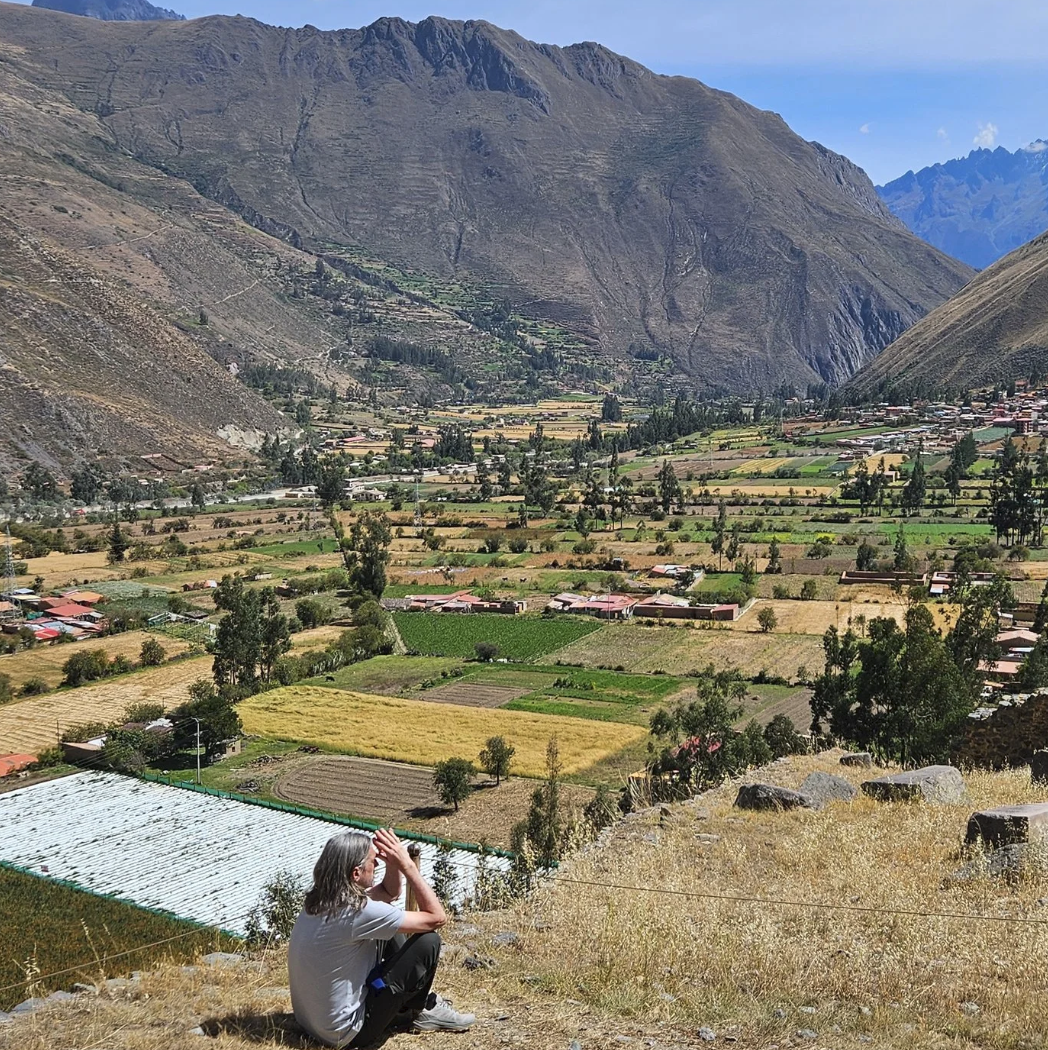 A person sitting on dry grass on a hillside, looking over a valley with farmland, houses, and mountains in the background.