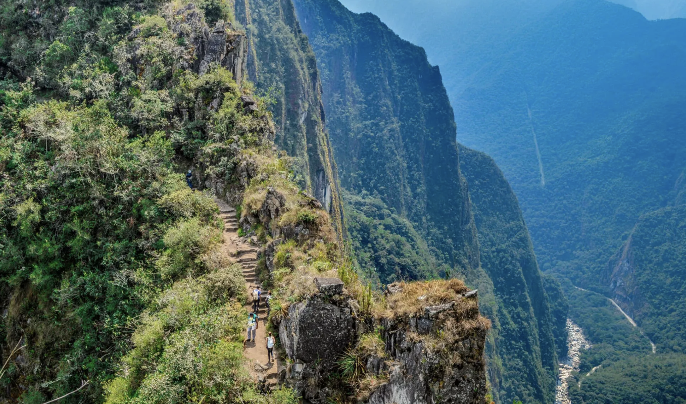 Trail with hikers along a steep mountain ridge with lush green vegetation and dramatic canyon views in the background.