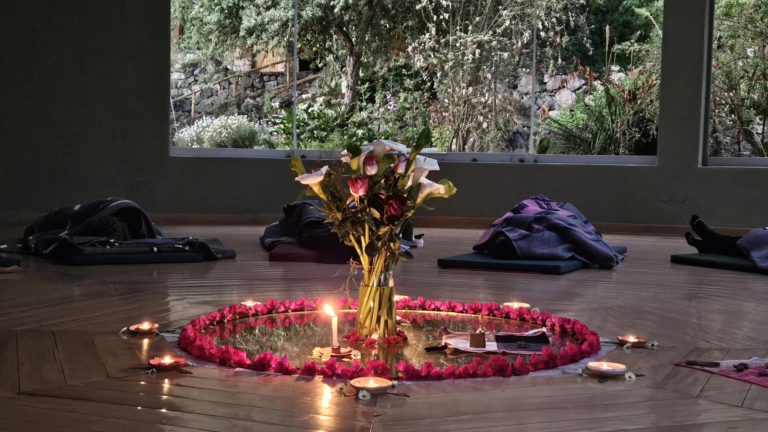 Indoor altar with flowers, candles, and personal items arranged in a circle on a wooden floor, overlooking a garden through large windows.
