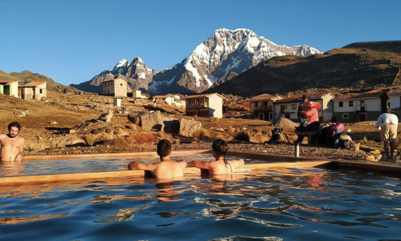 People enjoying hot spring in a mountainous landscape with snow-capped peaks and houses nearby.