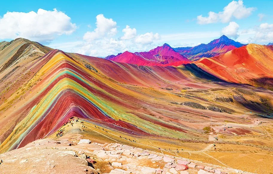 Panoramic view of Rainbow Mountain with colorful, layered peaks under blue sky with clouds.