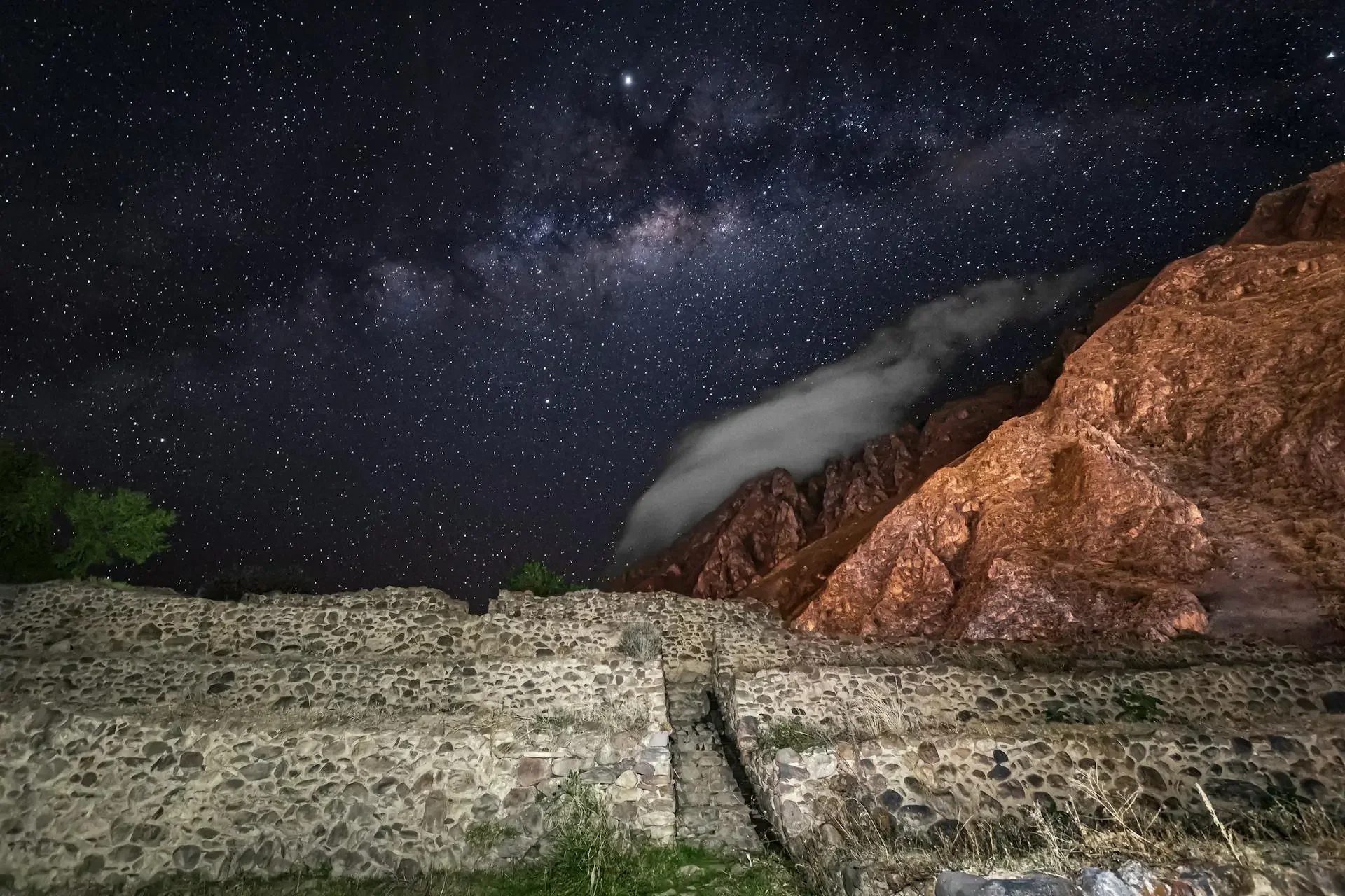 Night sky filled with stars above rocky terrain, with some greenery and a stone wall in the foreground.
