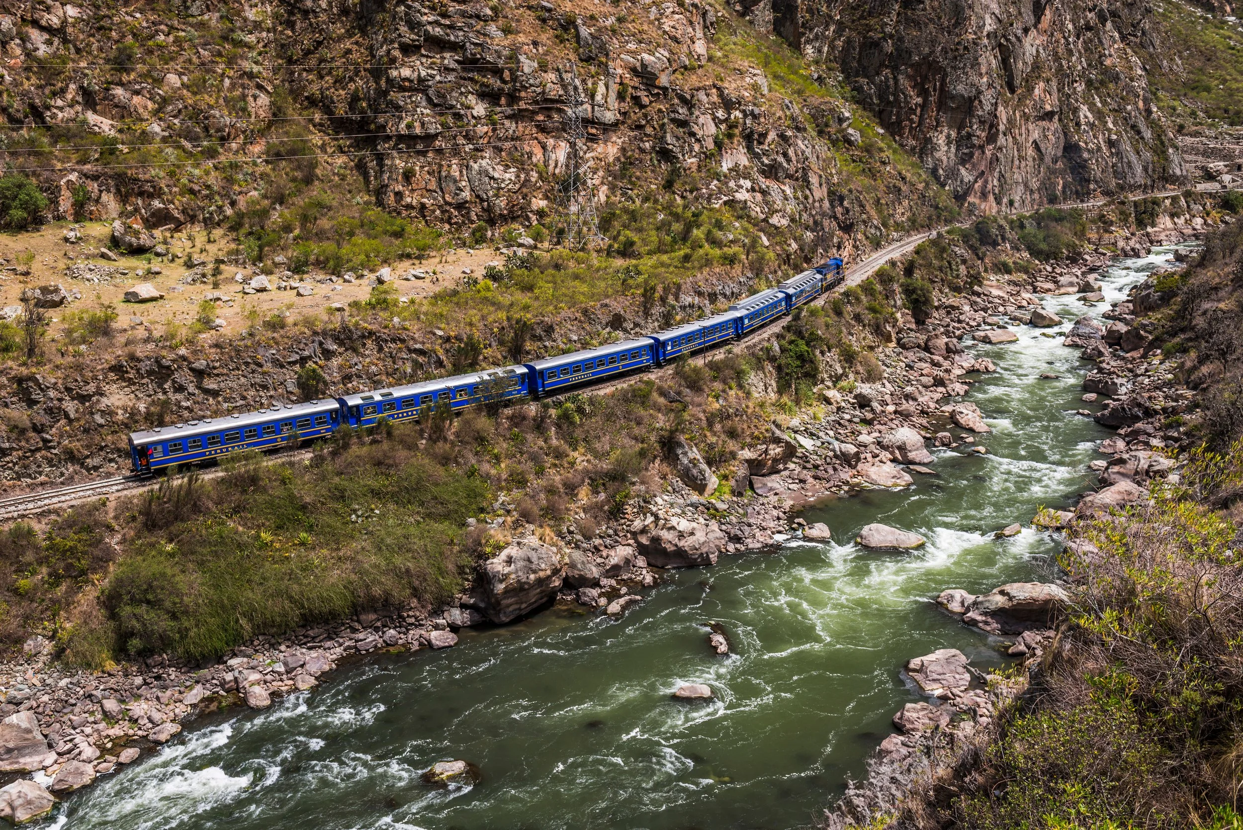 A blue passenger train traveling through a rocky canyon with a green river flowing alongside.