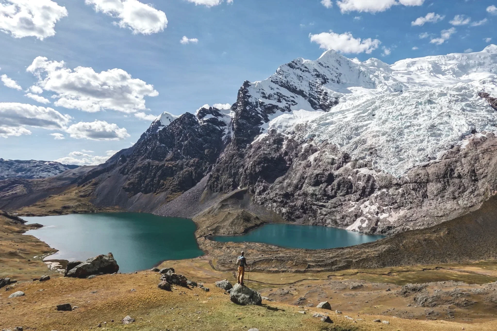 A hiker stands on a rock overlooking two turquoise mountain lakes with snow-capped peaks in the background and a partly cloudy sky.