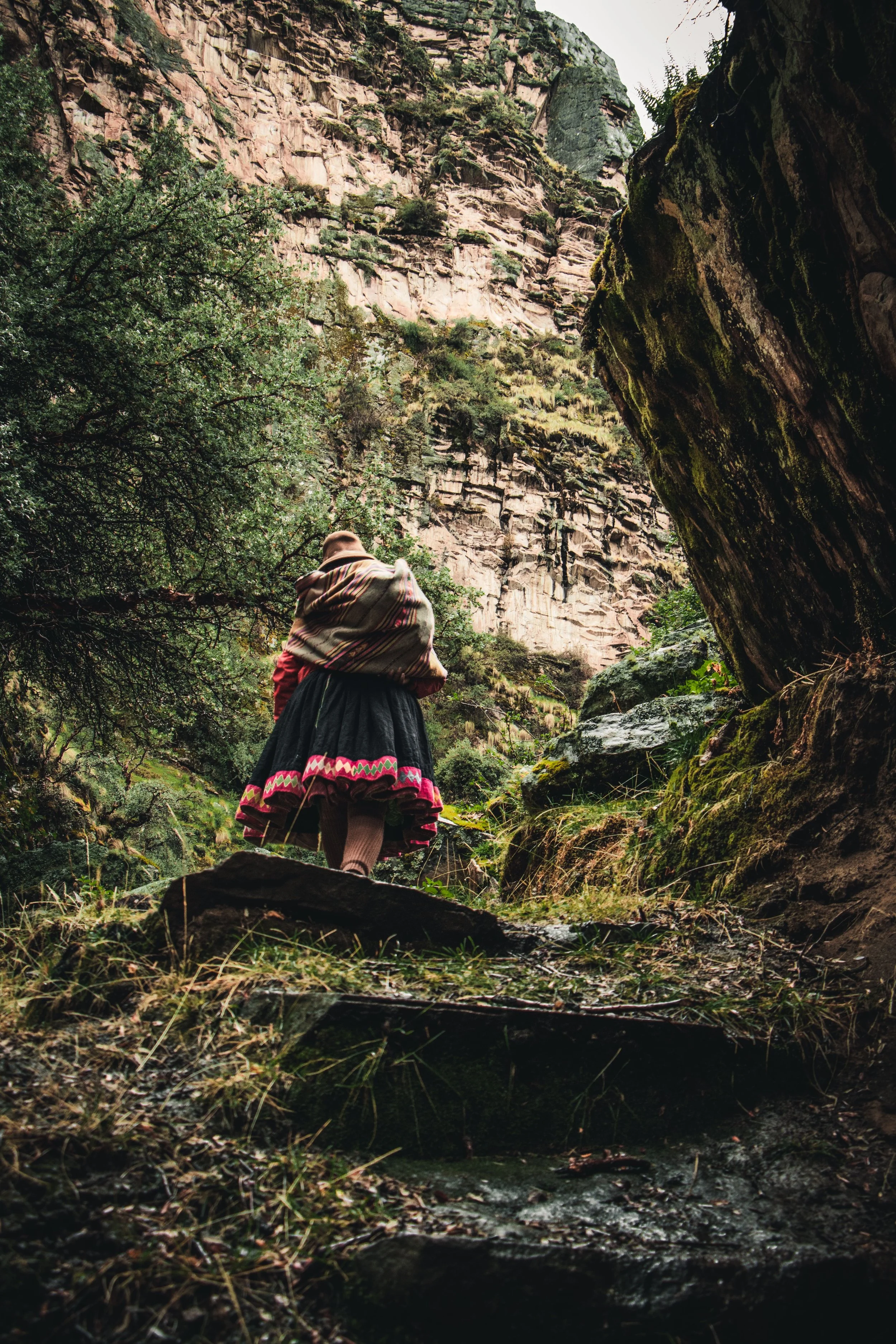 A person wearing a traditional woven shawl and skirt stands in a lush green canyon surrounded by tall moss-covered rocks and dense trees, overlooking a small stream.