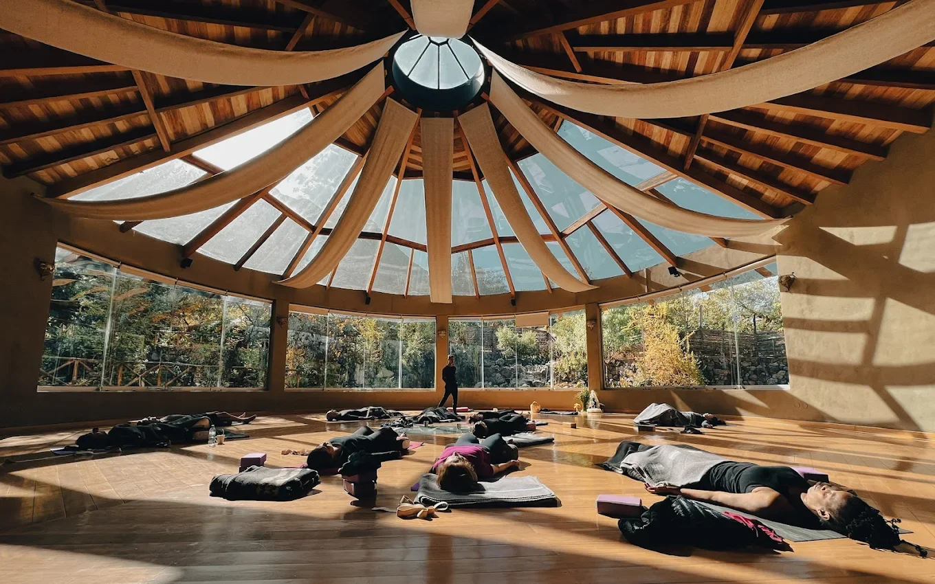 People participating in a yoga or meditation class in a spacious studio with large windows and a wooden ceiling with fabric drapes.