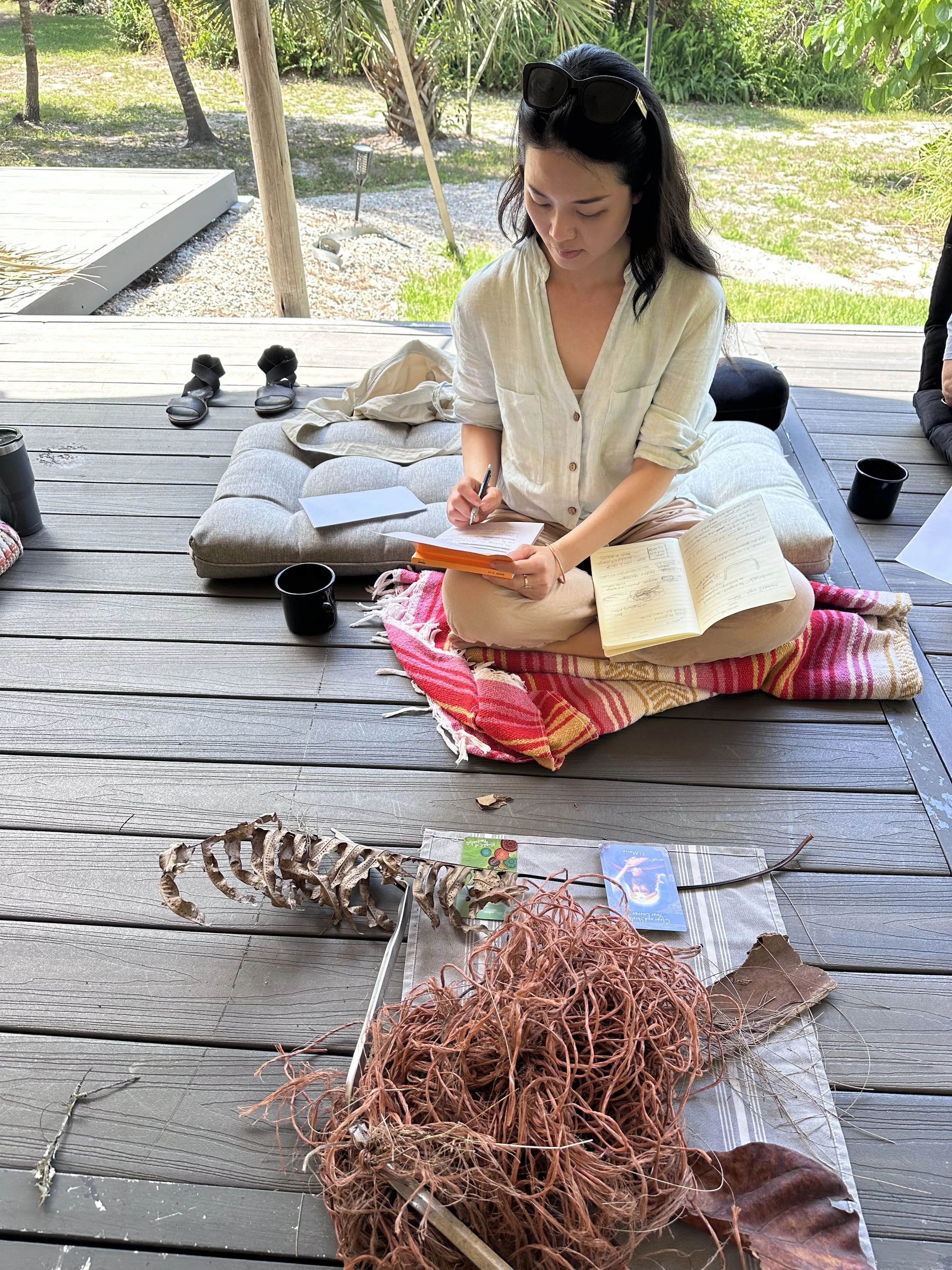 A woman sitting on a cushion on a wooden porch, writing in a notebook with another open notebook beside her. She has black hair, sunglasses on her head, and is wearing a beige blouse. In front of her are skeleton remains, including a ribcage and a spine, laid out on a cloth, with a couple of cards and leaves nearby. There are cups and personal items around her.