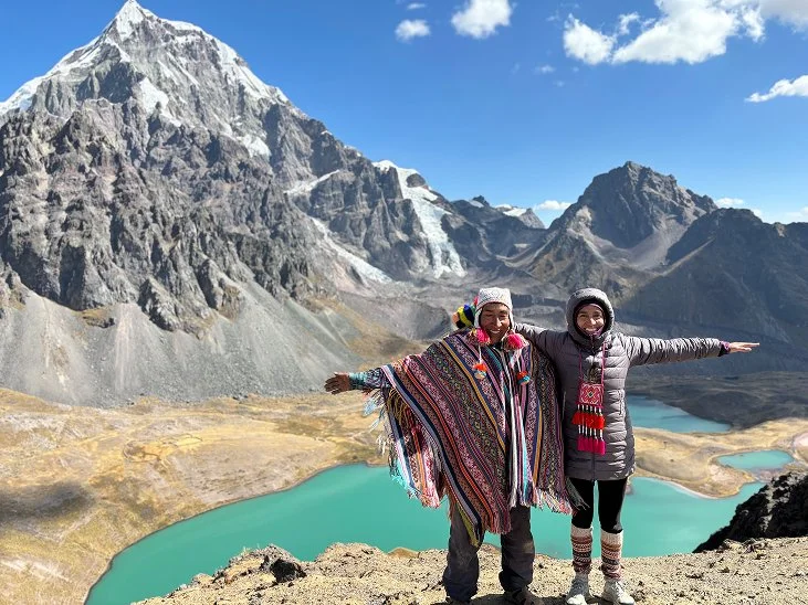 Two women in traditional Andean clothing standing on a mountain ledge with arms outstretched, overlooking turquoise lakes and rugged mountains with snow in the background, under a partly cloudy sky.
