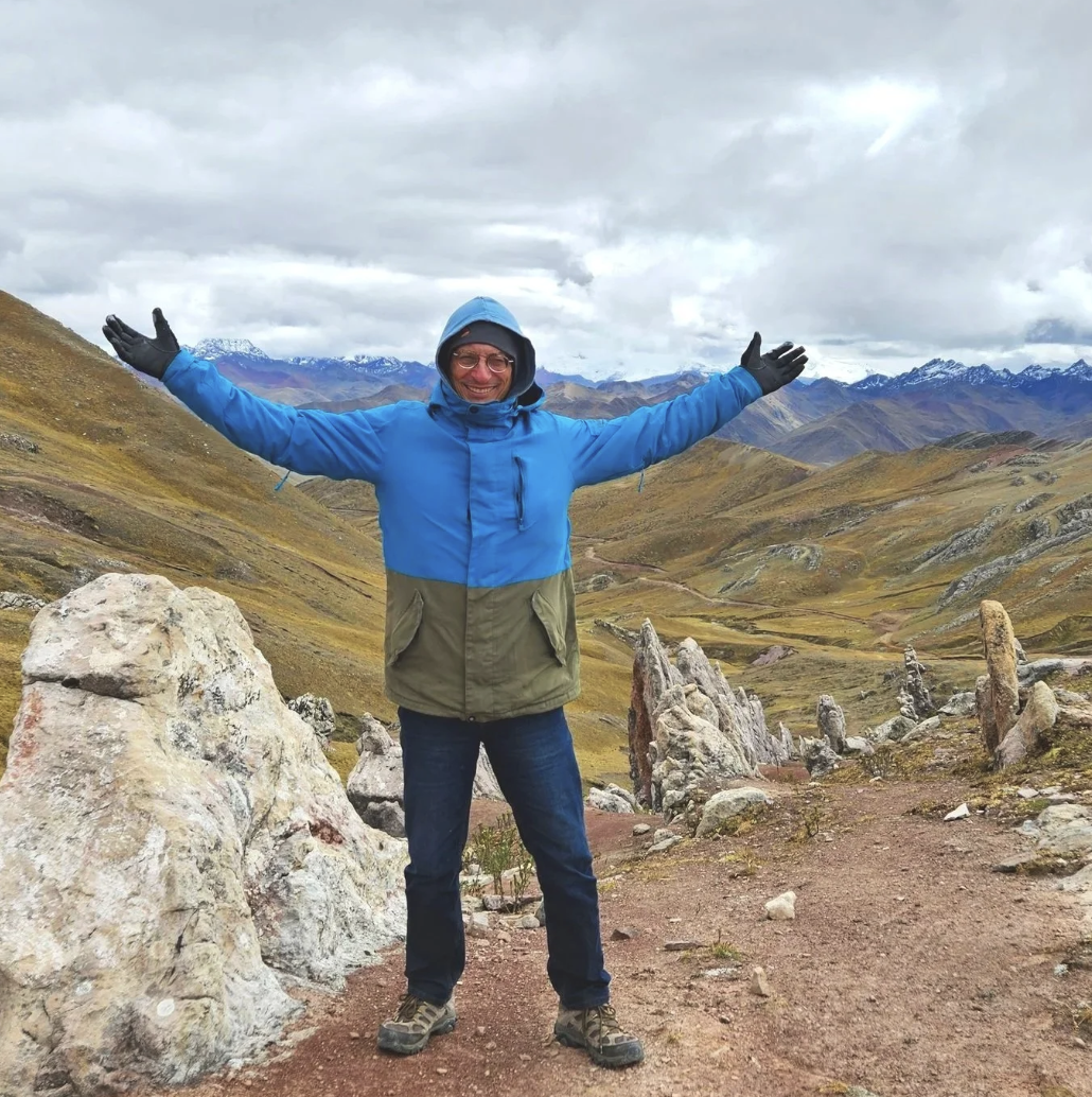 Man in blue and green jacket with black gloves and hiking boots standing on rocky trail in mountainous landscape with rolling hills, rocks, and distant snow-capped mountains under cloudy sky