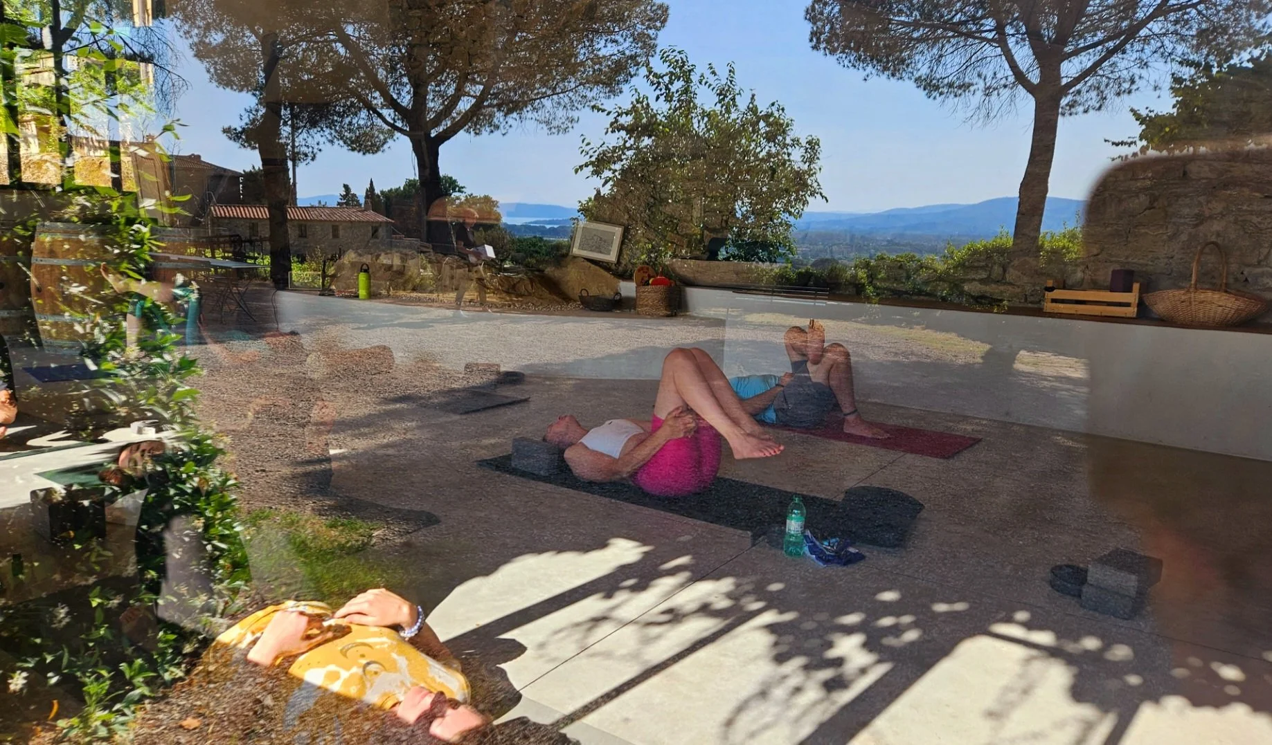 People practicing yoga indoors, viewed through a glass window with outdoor scenery and trees in the background.
