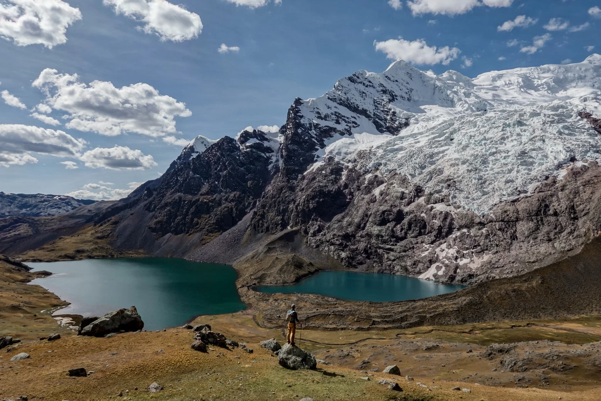 A hiker standing on a rock overlooking a mountain landscape with two green lakes, snow-capped peaks, and a partly cloudy sky.