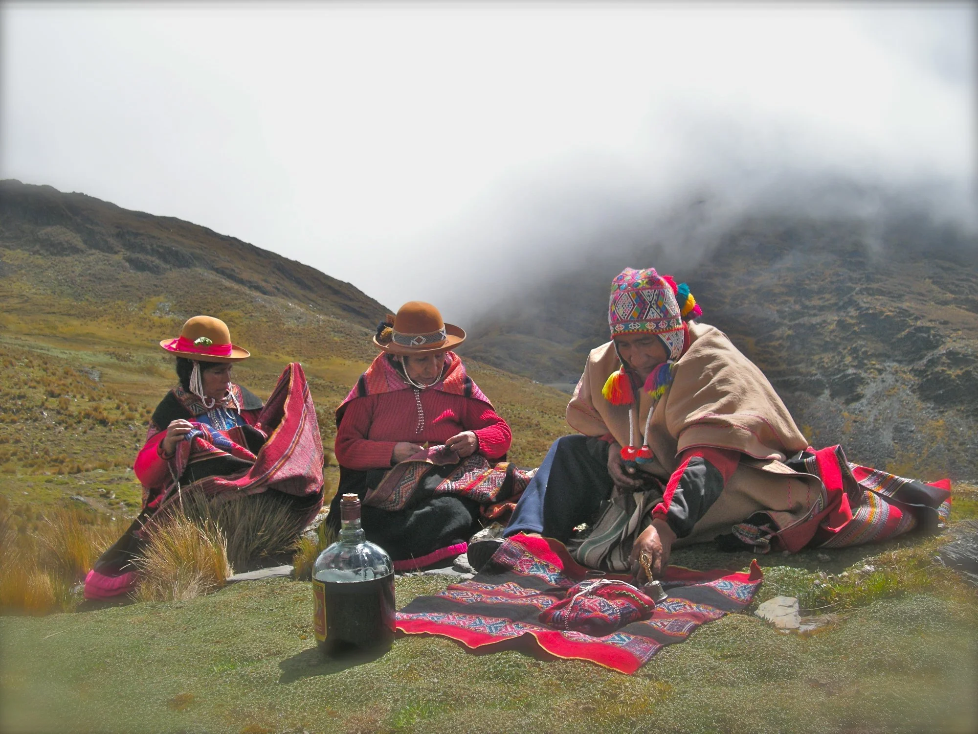 Three indigenous people dressed in traditional Andean clothing sit outdoors on a grassy area with mountains and clouds in the background.