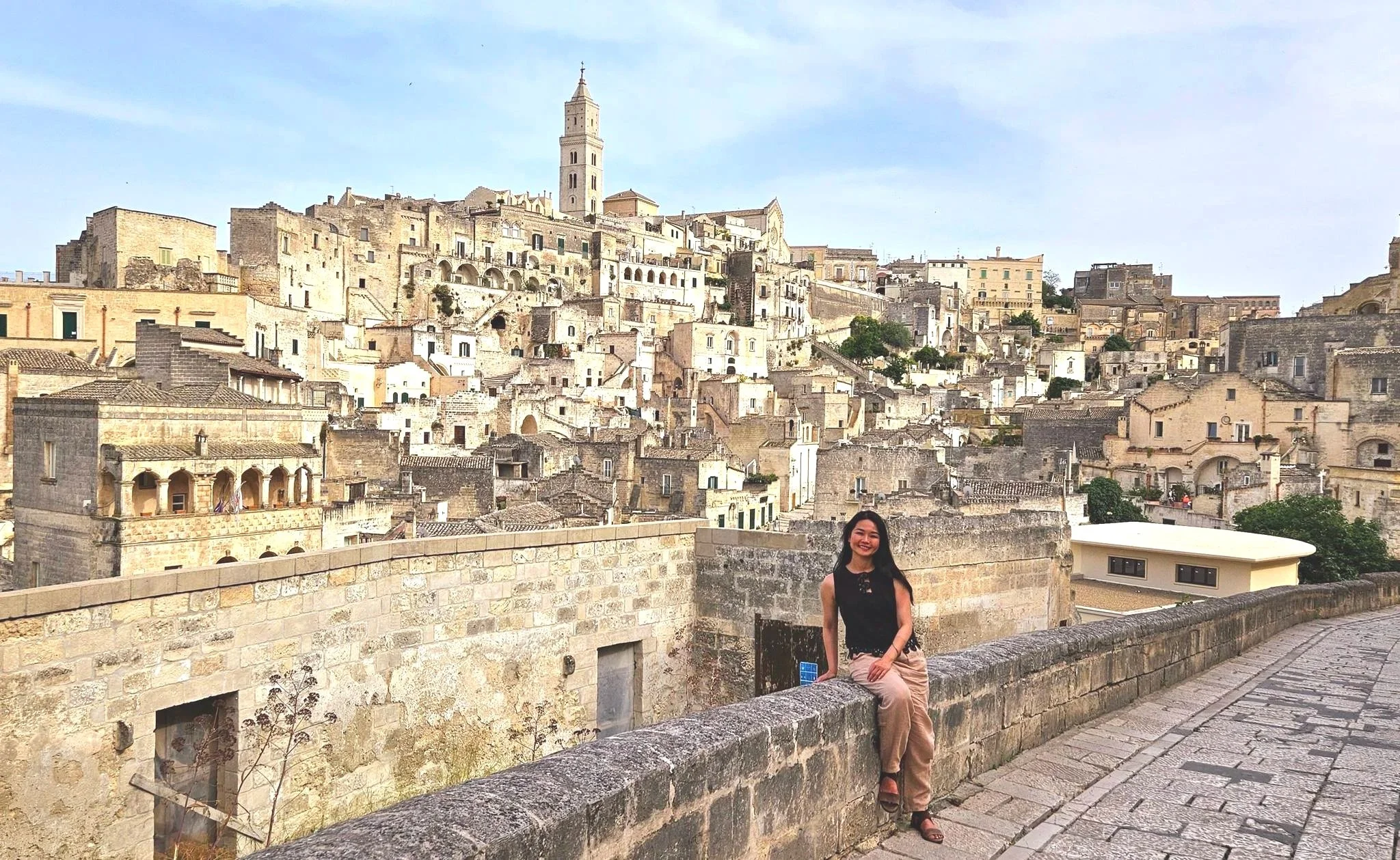 A woman sitting on a stone wall with an old European town with stone buildings, narrow streets, and a church steeple in the background.