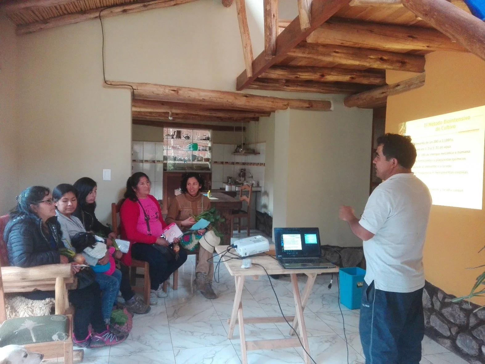 A man giving a presentation to a group of women in a cozy, rustic room with wooden ceiling beams. The women are seated, listening attentively, and some hold papers or notebooks. There is a projection screen displaying a slide in Spanish titled 'El Metodo Biointensivo de Cultivo' and a laptop connected to a projector on a wooden table.