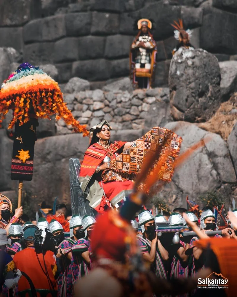 A cultural festival with women dressed in traditional attire, carrying colorful umbrellas, and a woman in a red dress sitting on a decorated palanquin, surrounded by soldiers in uniform wearing silver helmets and black masks, in front of a rocky landscape.