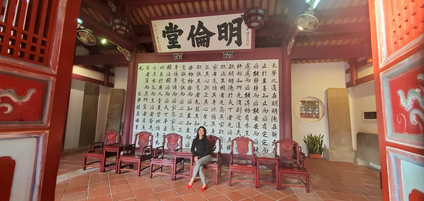 A woman sitting on a wooden chair in a traditional Chinese hall with red furniture and a large calligraphy scroll on the wall behind her.