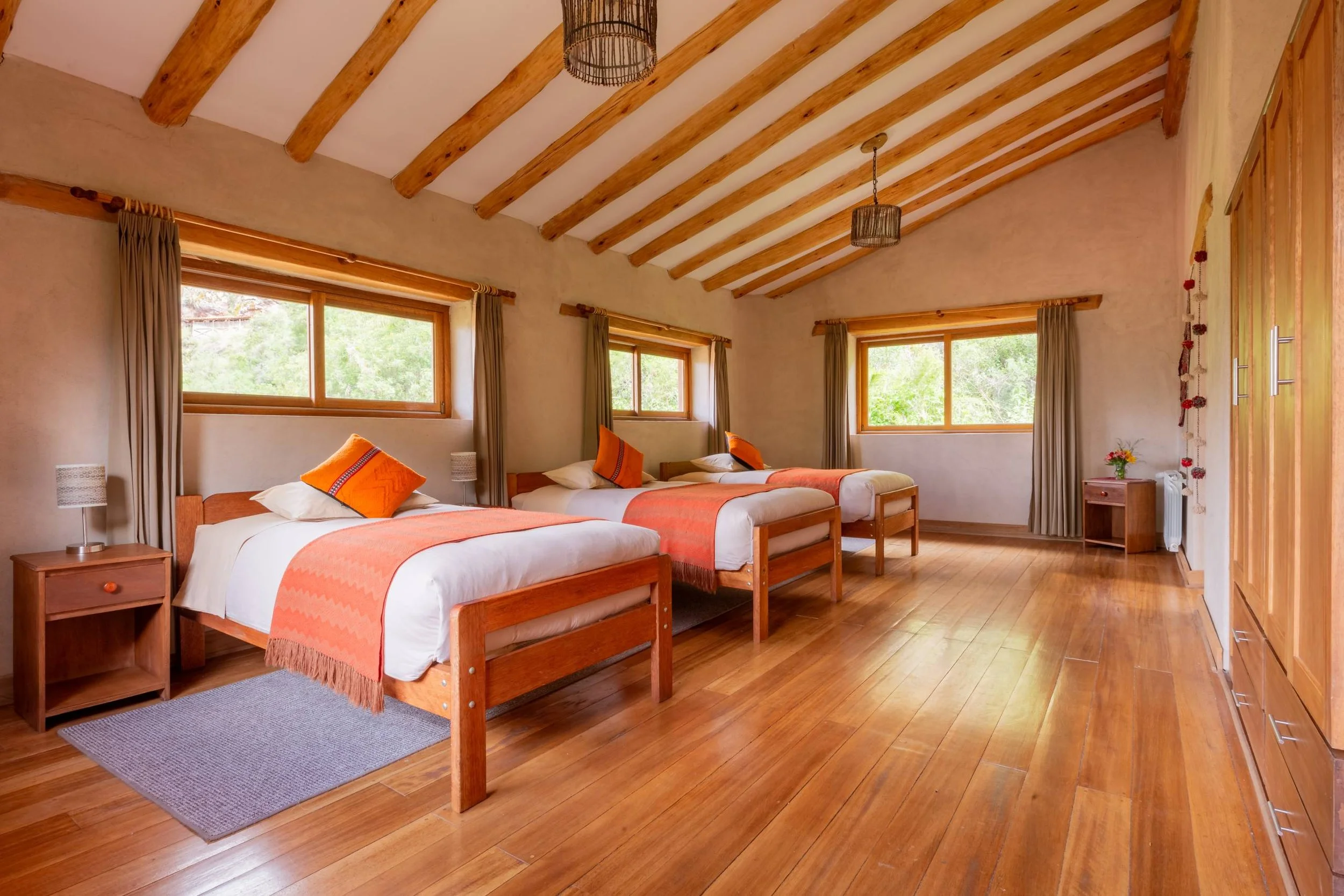 A spacious bedroom with three single beds with orange and white bedding, wooden nightstands with lamps, hardwood flooring, large windows with brown curtains, and a wooden wardrobe in the corner.
