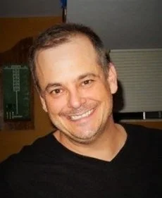 A smiling man with short, dark hair and a beard, wearing a black shirt, indoors near some windows.