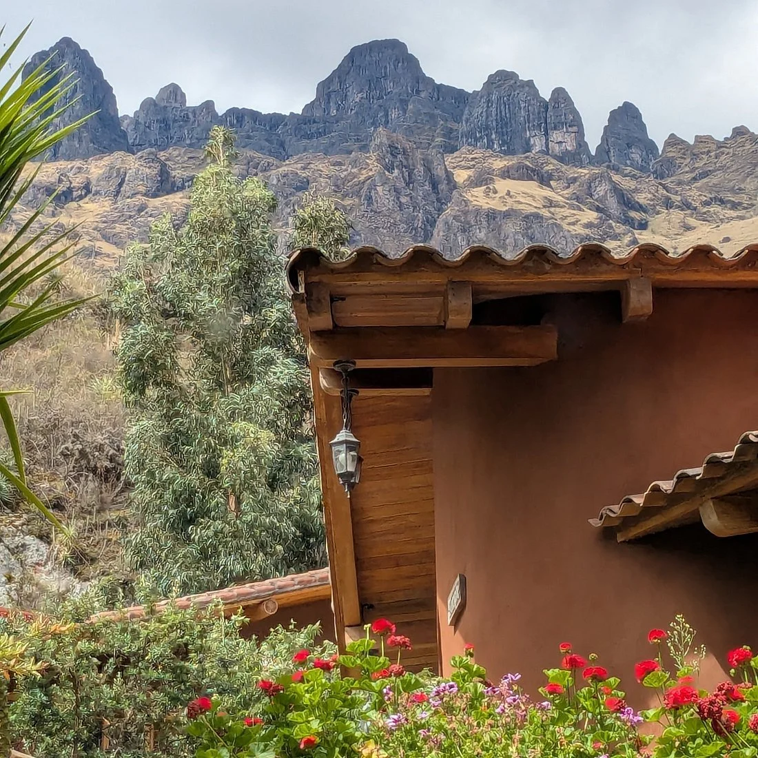 A wooden house with a tile roof, surrounded by colorful flowers and greenery, with mountains in the background.