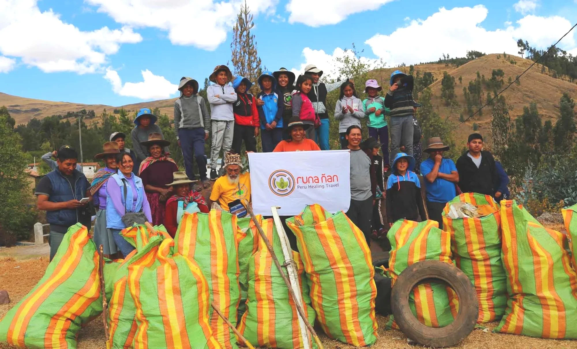 Group of people in outdoor rural setting holding a banner with a logo and text, surrounded by colorful striped bags, with hills and trees in the background.