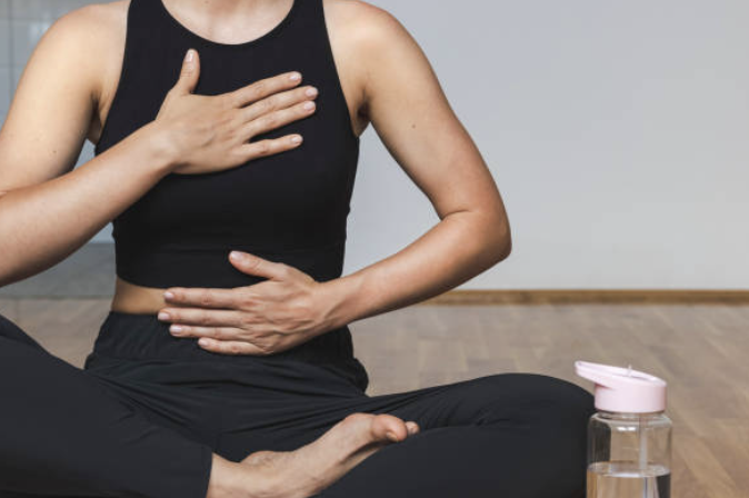 A person is practicing meditation or yoga, sitting cross-legged on the floor, with one hand on their chest and the other on their stomach, and a water bottle nearby.