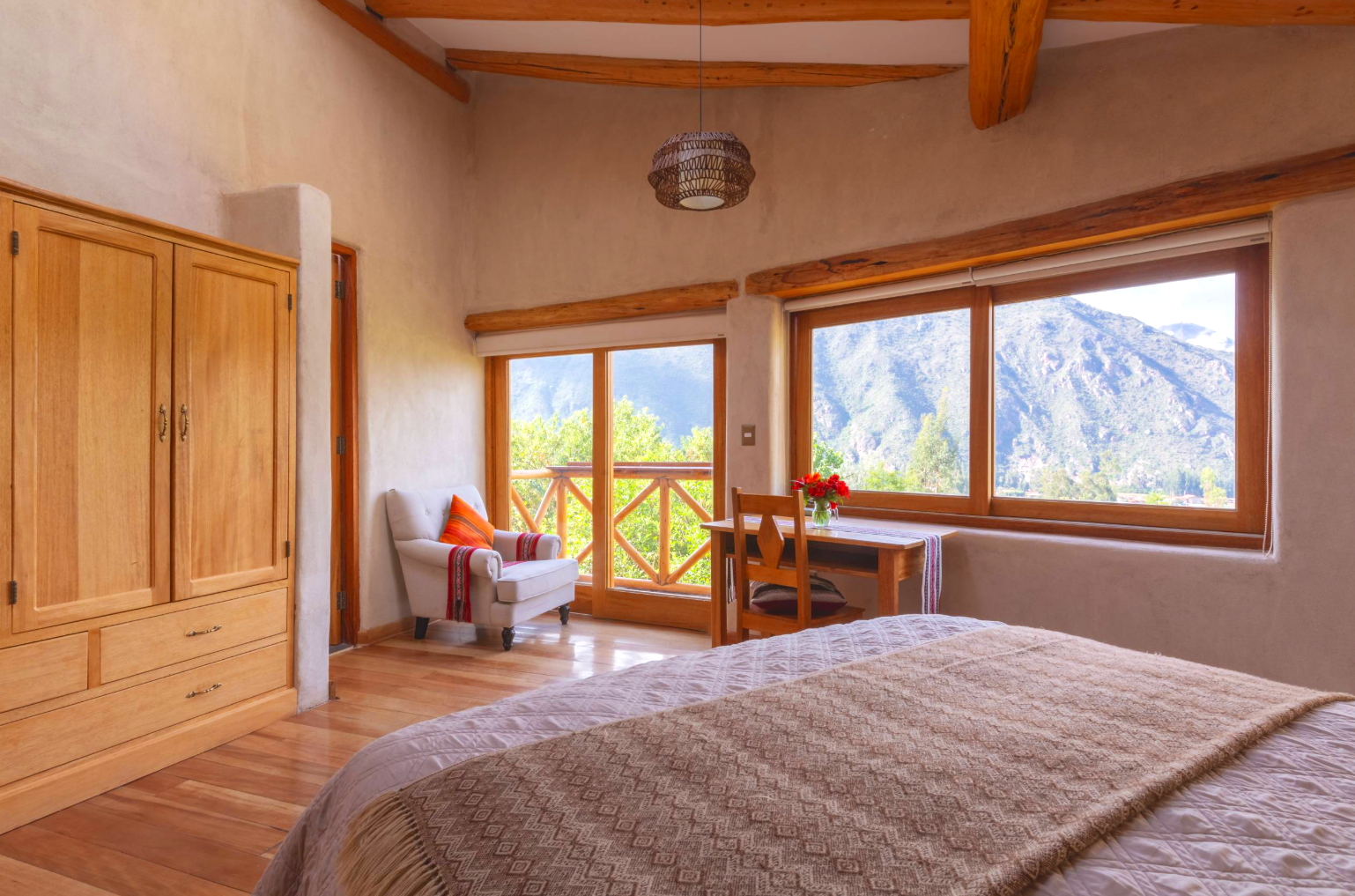 Bedroom with a large window view of mountains, wooden furniture, a white armchair with an orange pillow, a wooden desk with a chair and a vase of red flowers, a bed with a beige quilt, and wooden flooring and ceiling beams.