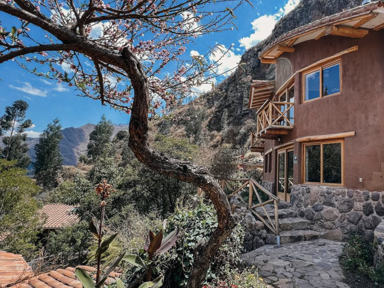 A house with rounded walls and large windows located among trees and mountains, with a stone pathway and wooden railings.