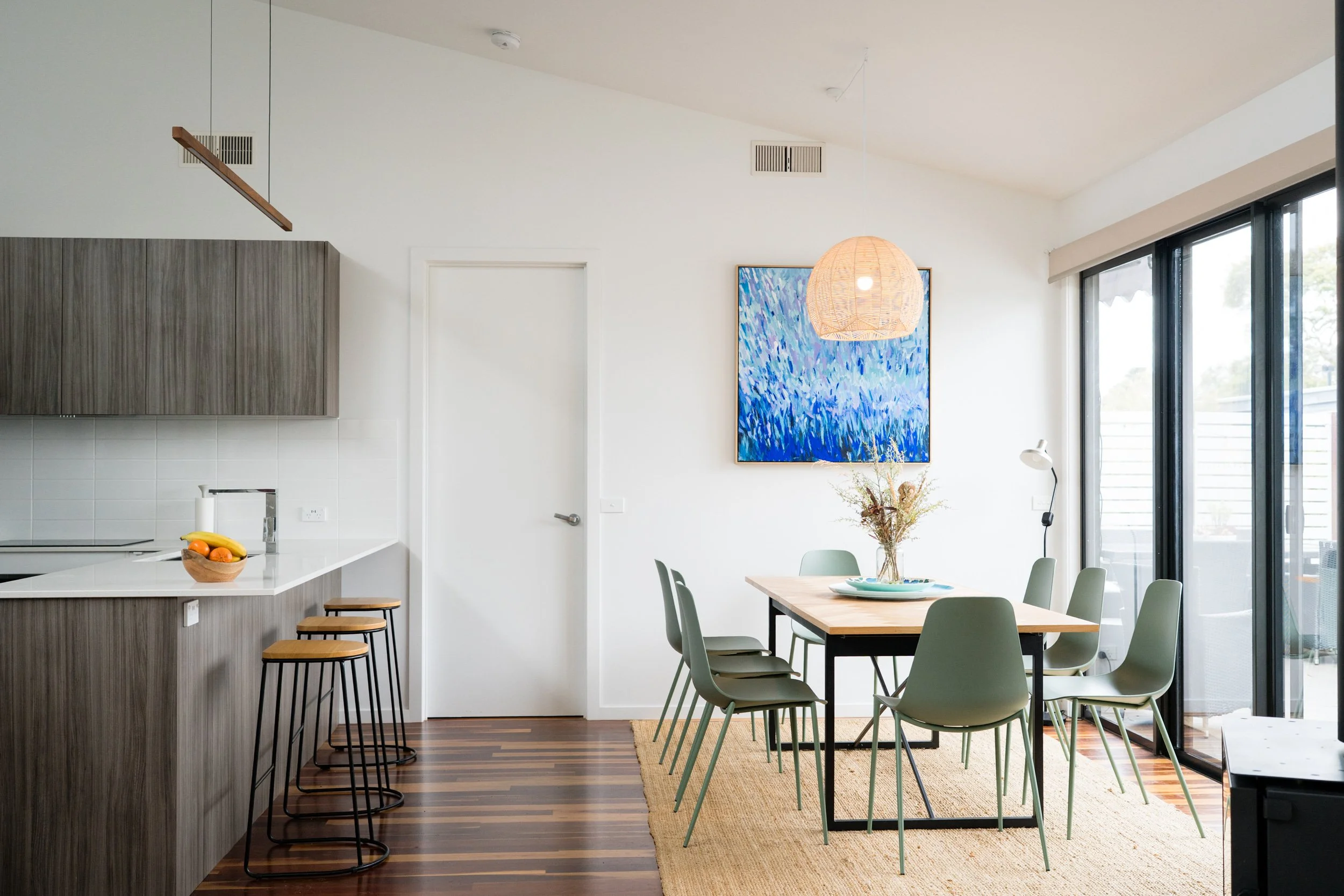 Modern dining area with six green chairs around a rectangular wooden table, a vase with dried flowers, a large abstract blue painting on white wall, a pendant light, and sliding glass door leading outside. Kitchen counter with fruit bowl in the foreg
