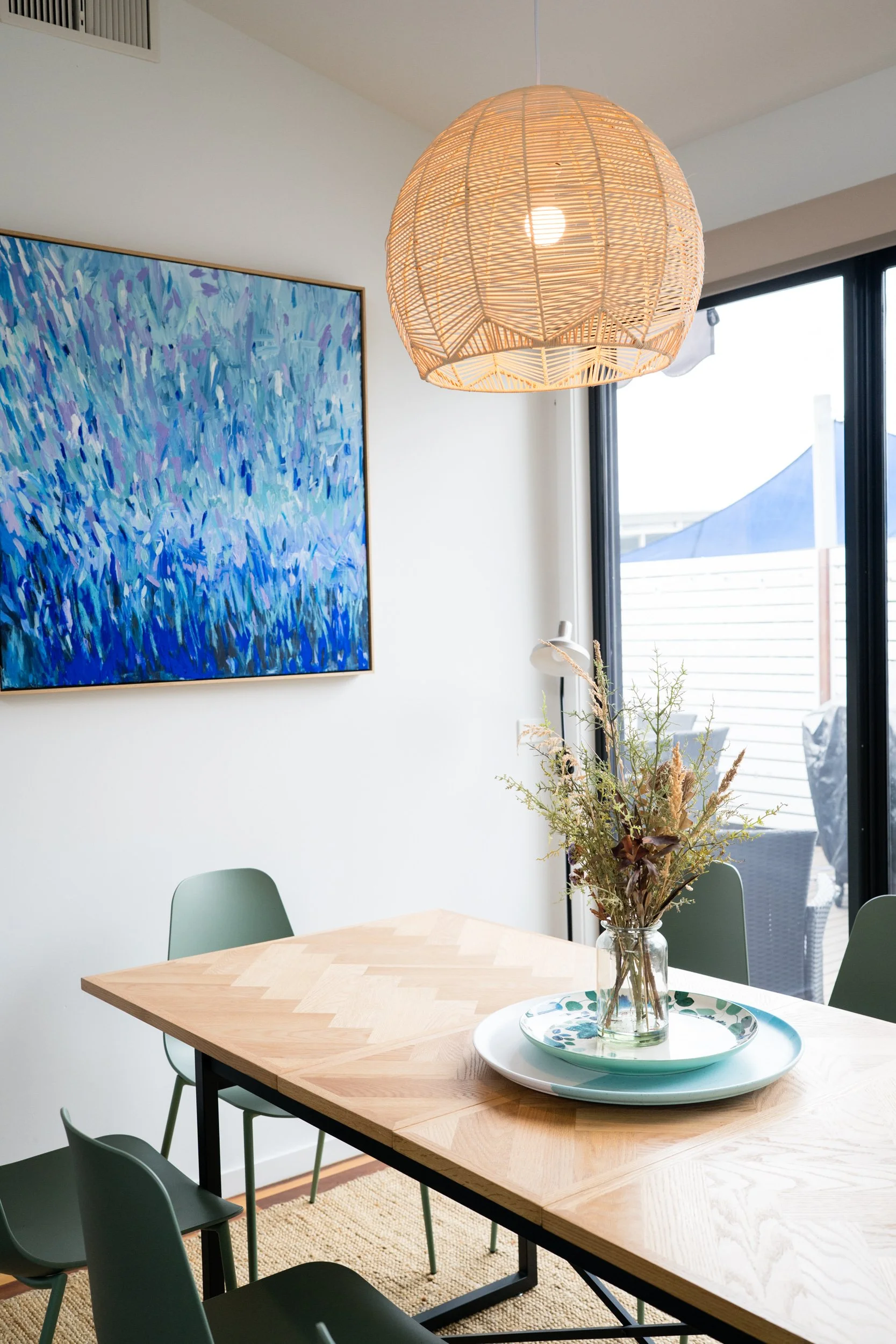 A dining room with a wooden table, green chairs, a glass vase with dried flowers, a colorful abstract painting on the white wall, a rattan pendant light, and a large window letting in natural light.