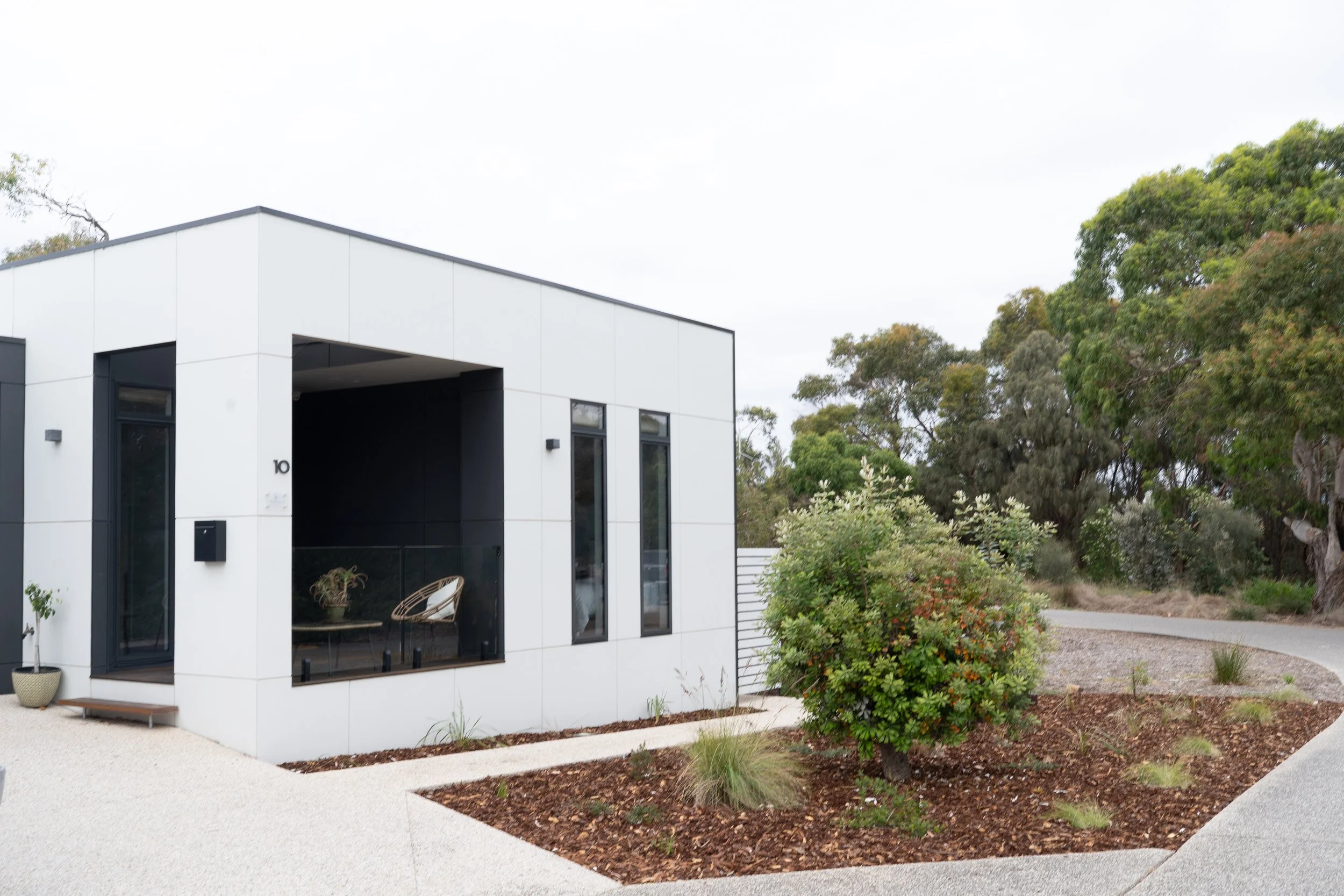 Modern eco white house with large glass windows, black accents, small front yard with shrubs and a curved pathway, trees in background.
