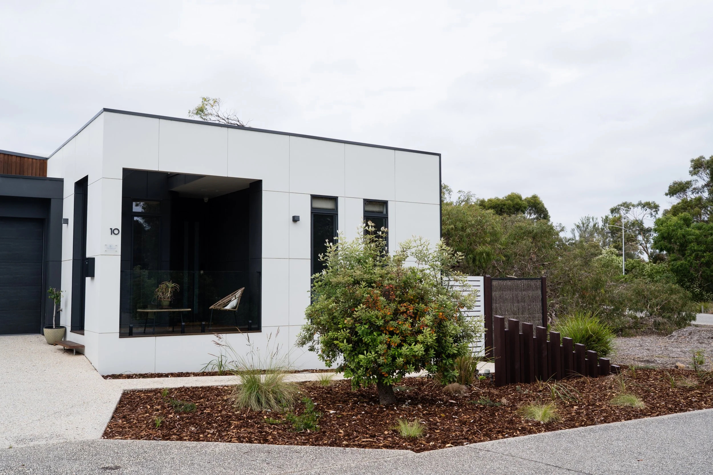 Modern white house with black accents, featuring a large front window with a small balcony, surrounded by plants and a landscaped yard.