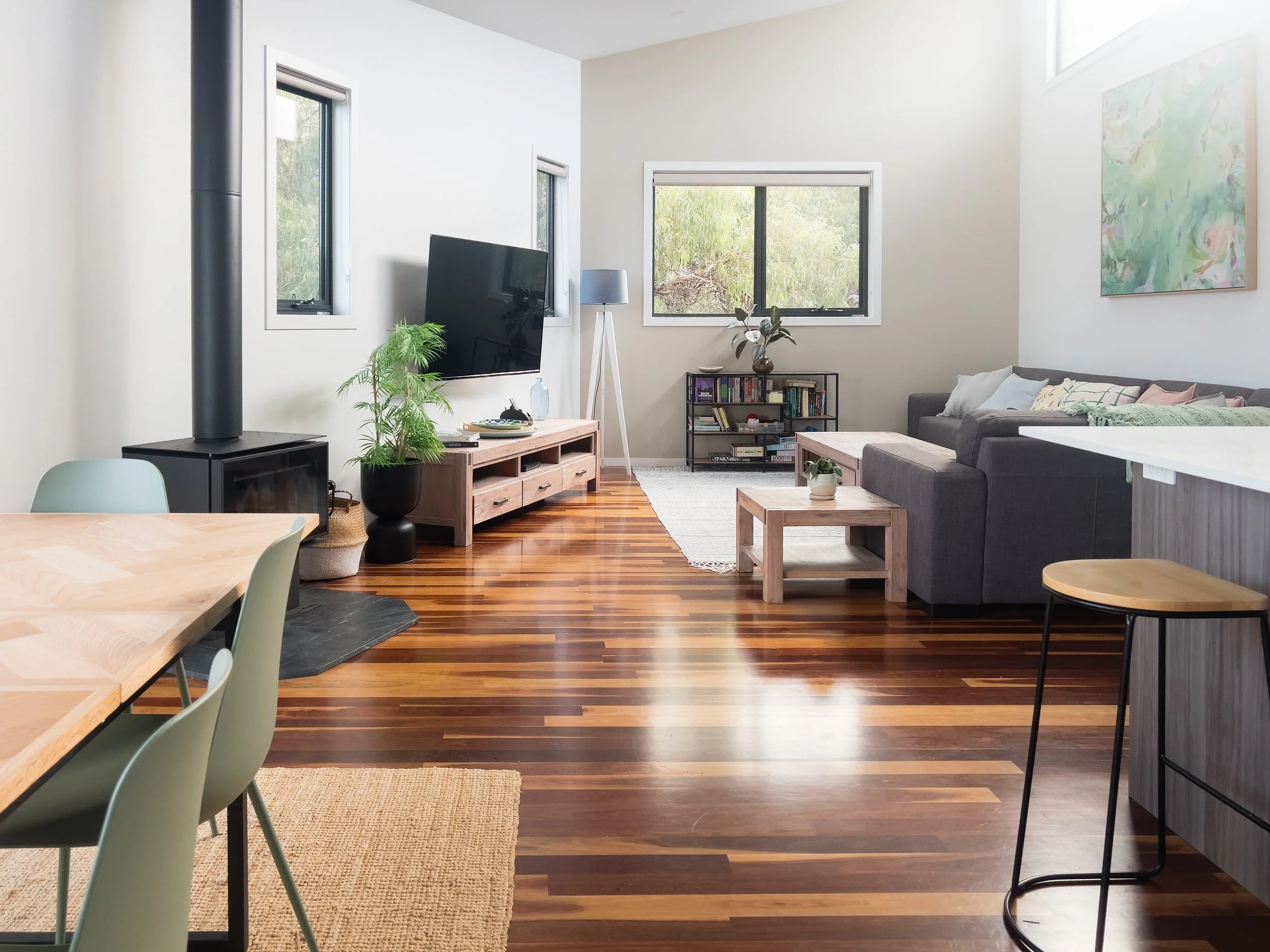 Open concept living room with hardwood floors, a gray sectional sofa, coffee tables, a TV on a stand, potted plants, windows, and artwork on the wall.