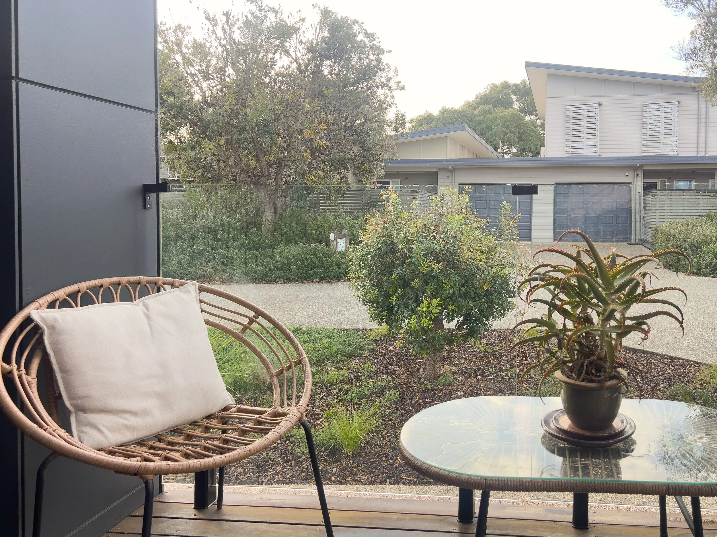 A cozy outdoor seating area with a rattan chair with a white pillow and a glass-top table with a potted succulent plant, overlooking a garden and neighboring houses through a glass window.