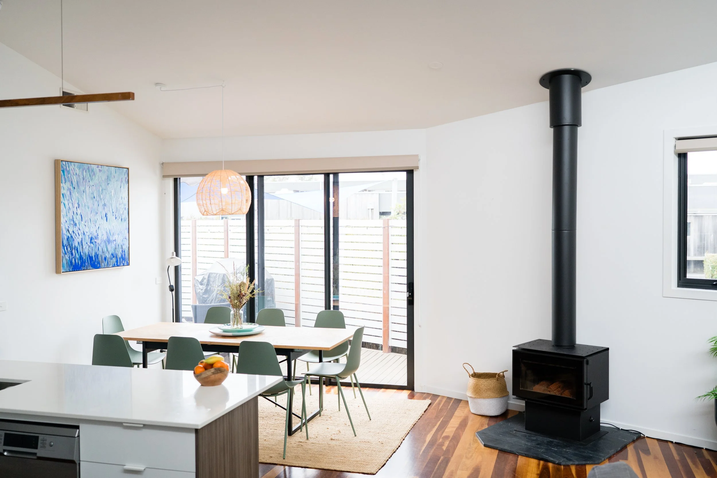Modern dining area with a wooden table and green chairs, sliding glass door leading outside, and a black wood stove in the corner.