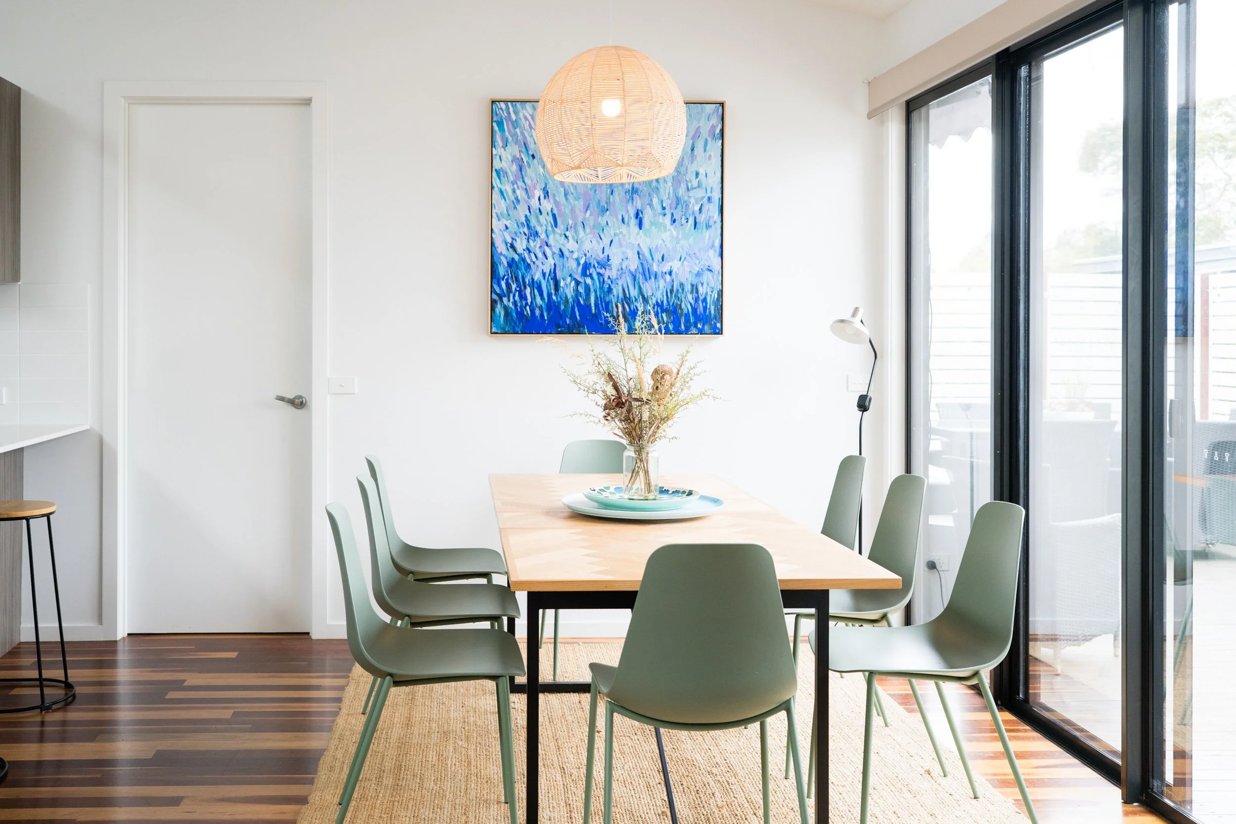 Modern dining room with a wooden table surrounded by eight green chairs, a vase with dried flowers, a large blue abstract painting, a rattan pendant light, and sliding glass doors leading to an outdoor deck.