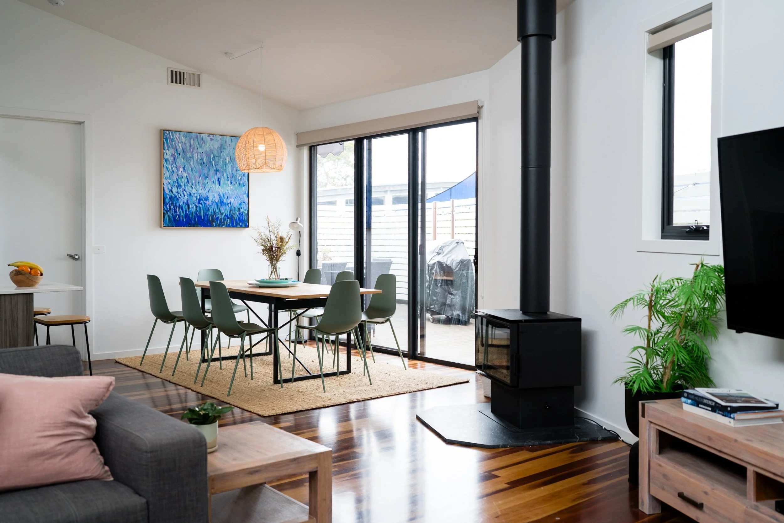 Modern living room with a dining area, wood floors, a black wood stove, large sliding glass door leading to a balcony, a small TV, a gray sofa with pink pillow, and a wooden coffee table with a plant.