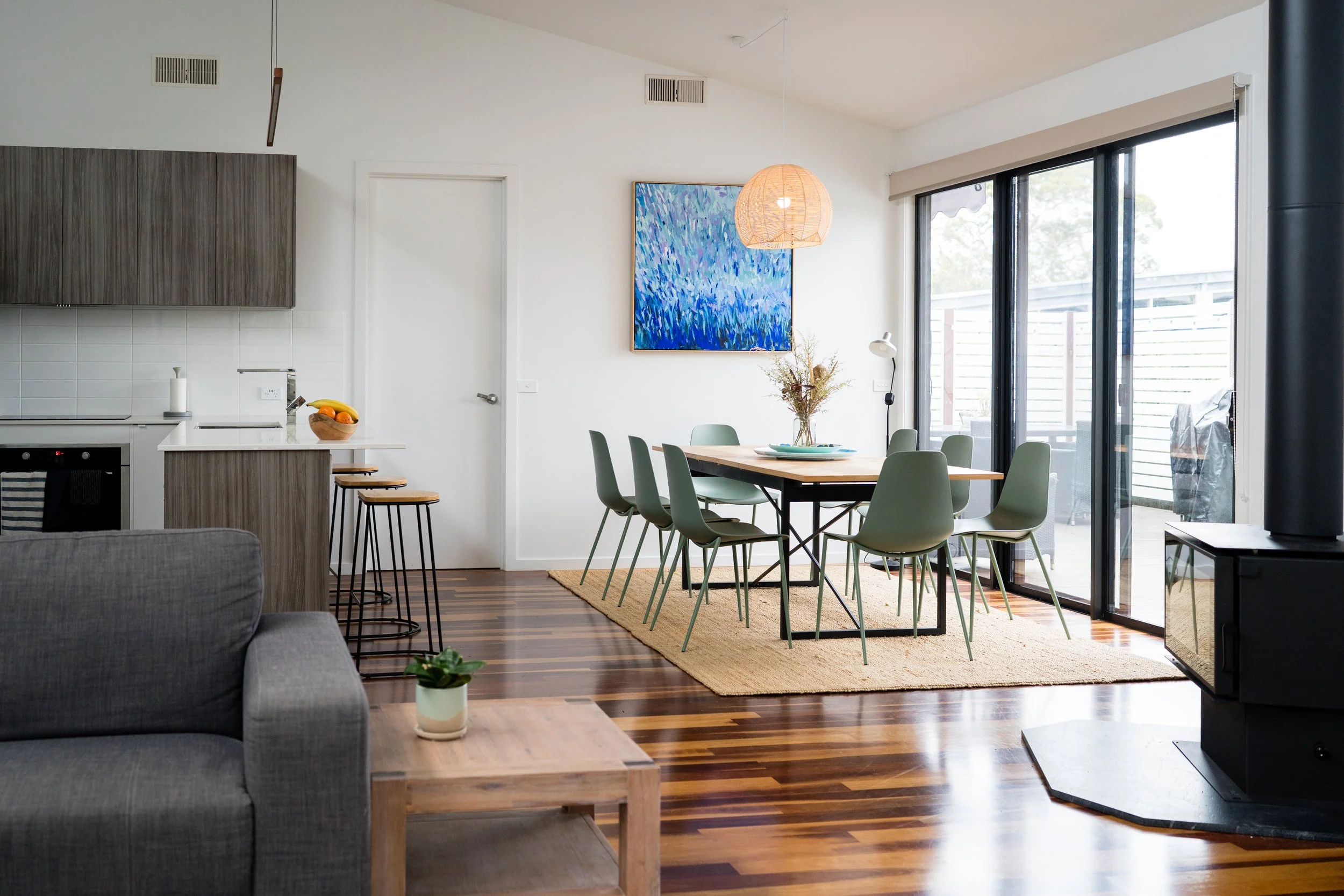Open-concept living and dining area with wooden floors, a gray couch, a small wooden table with a potted plant, a gray kitchen island with stools, a dining table with green chairs, a large sliding glass door, a colorful abstract painting, and a fireplace.